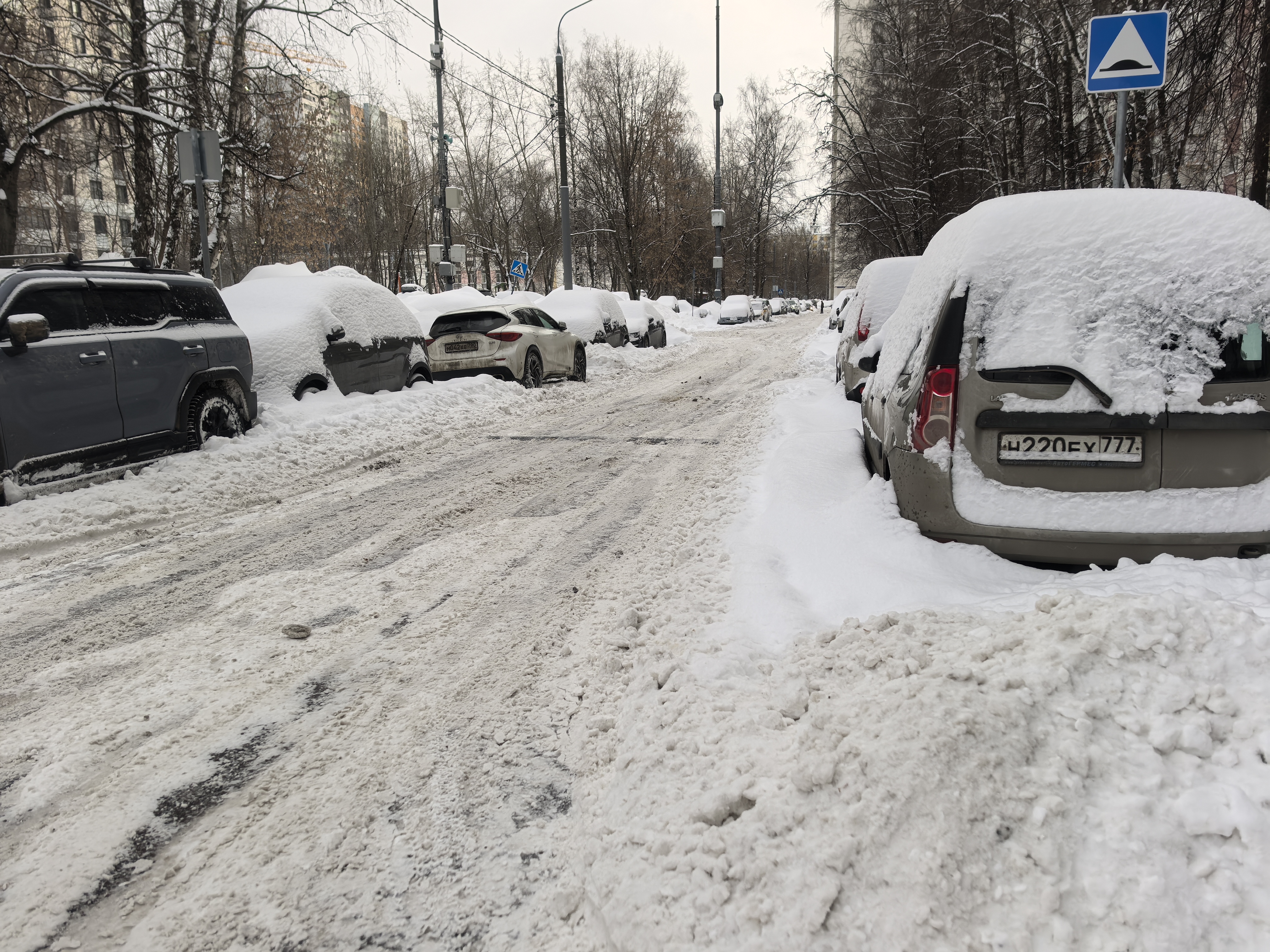 Проезжая часть без пешеходного перехода, покрытая снегом и слякотью. С обеих сторон дороги припаркованы автомобили, частично занесенные снегом. Видимых тротуаров нет, пешеходы вынуждены идти по проезжей части.