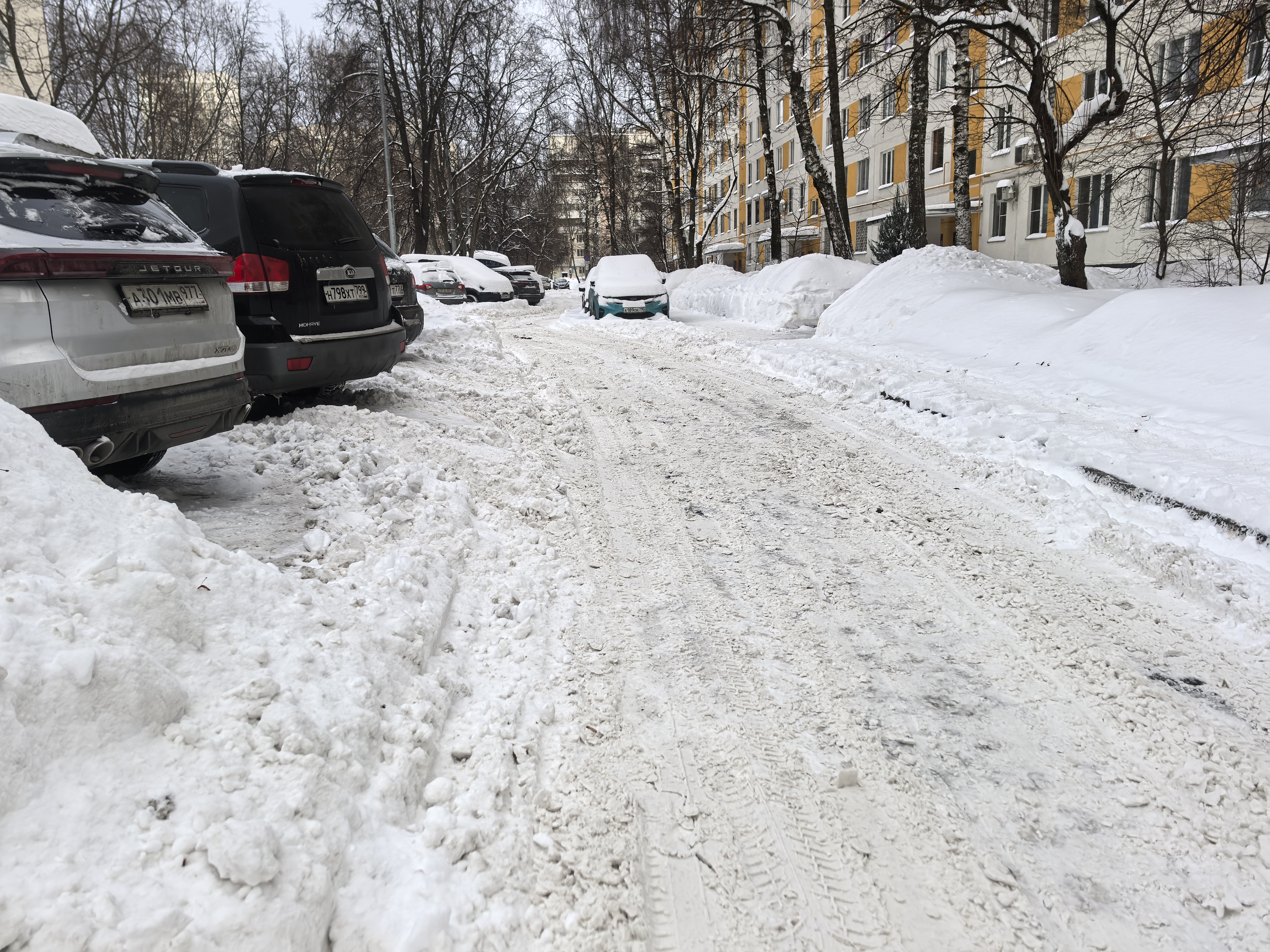 Дворовая территория с припаркованными автомобилями, покрытая снегом. Проезжая часть узкая, тротуары полностью засыпаны снегом.