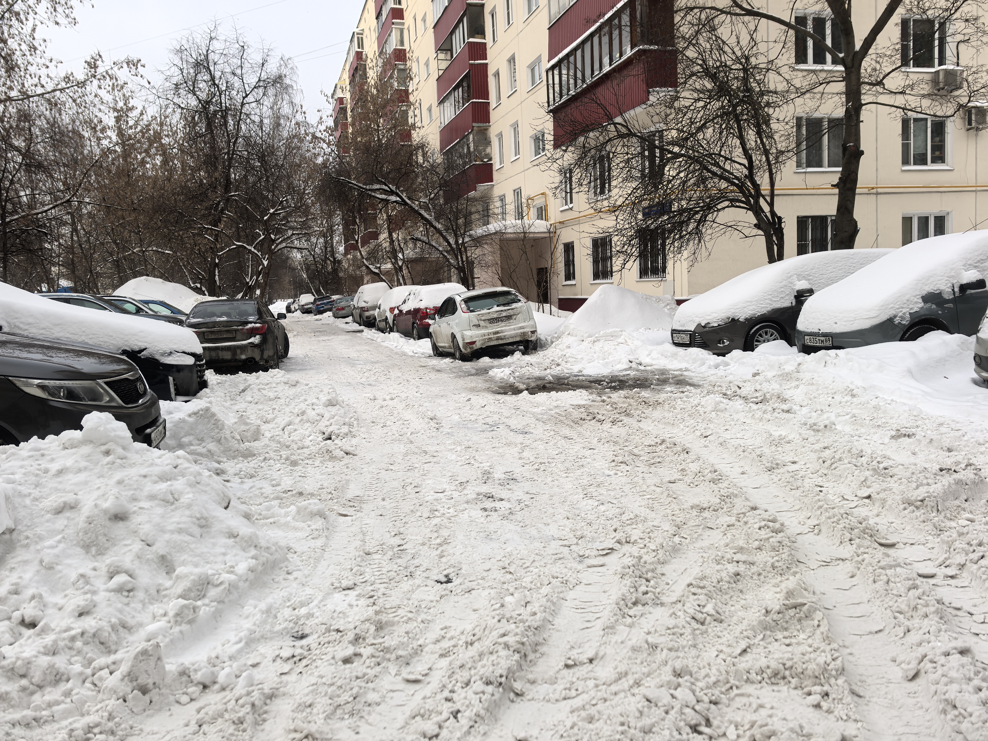Дворовая территория, проезд между жилыми домами, покрытый снегом, с припаркованными автомобилями по обеим сторонам. Пешеходных переходов и разметки нет.