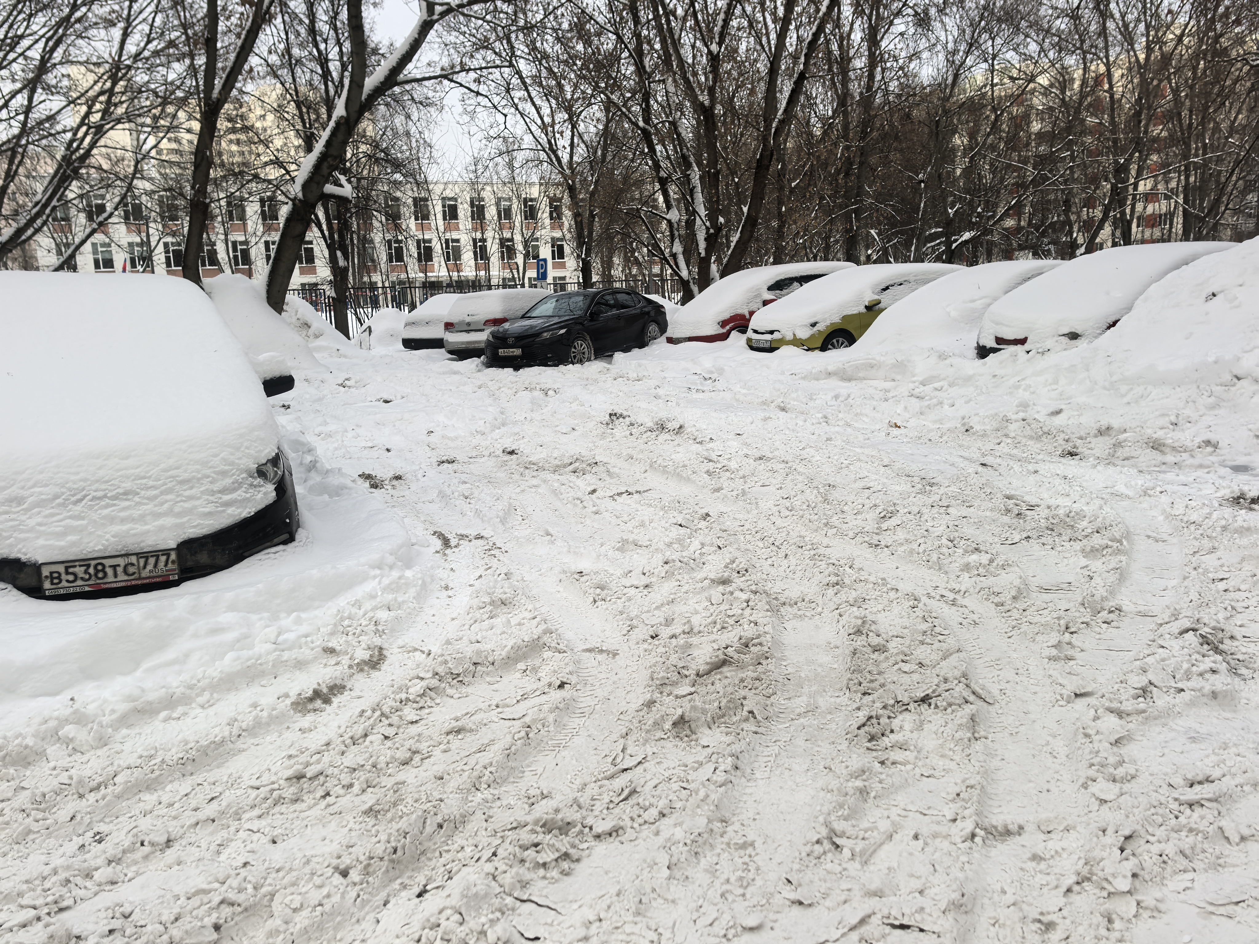 Дворовая территория, покрытая снегом, с припаркованными автомобилями. Видна проезжая часть, очищенная от снега, но покрытая следами шин. На заднем плане видны жилые дома.
