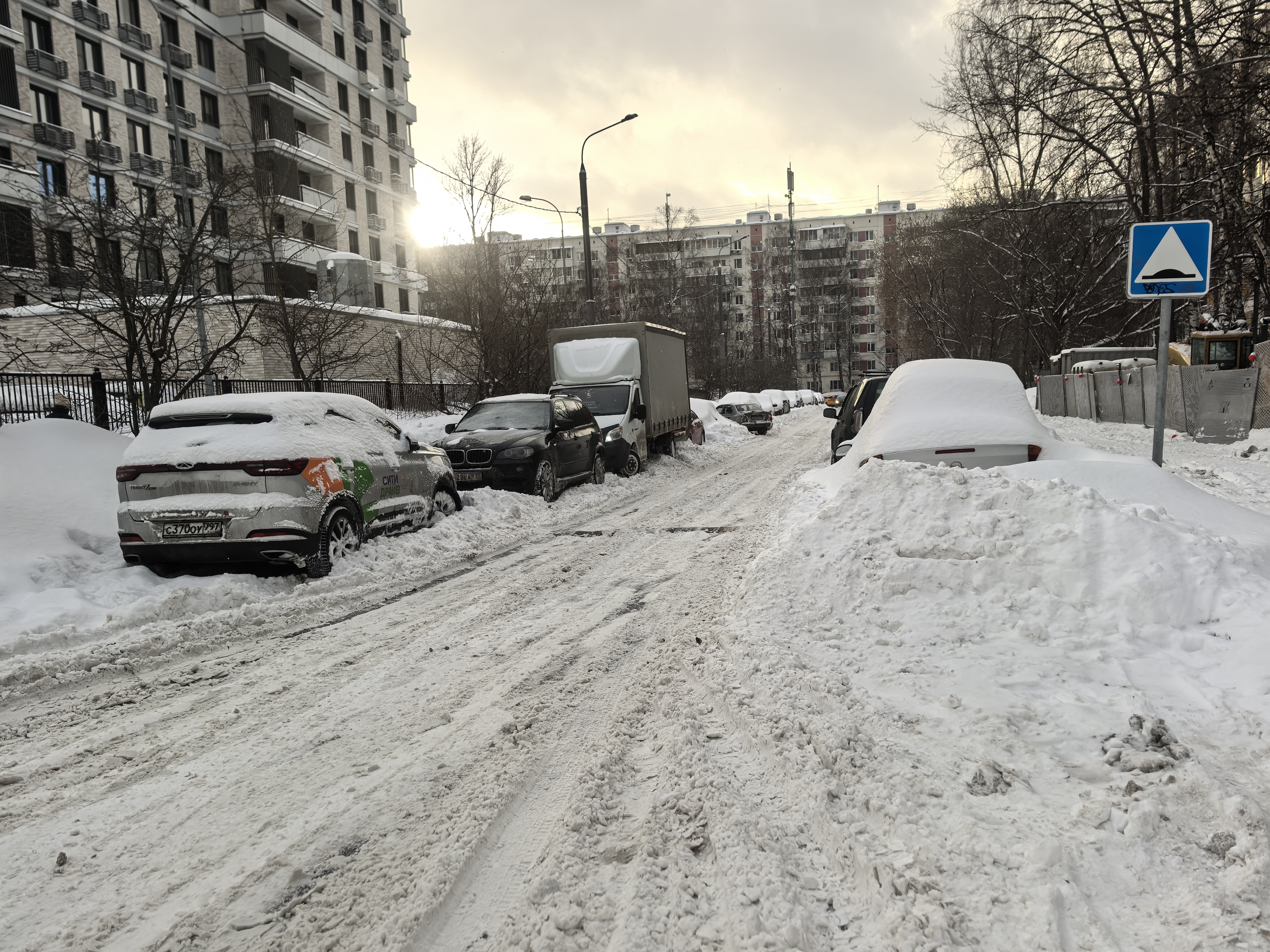 Участок дороги без пешеходного перехода, покрытый снегом и грязью. С обеих сторон дороги припаркованы автомобили, частично занесенные снегом. Видны жилые дома и деревья.