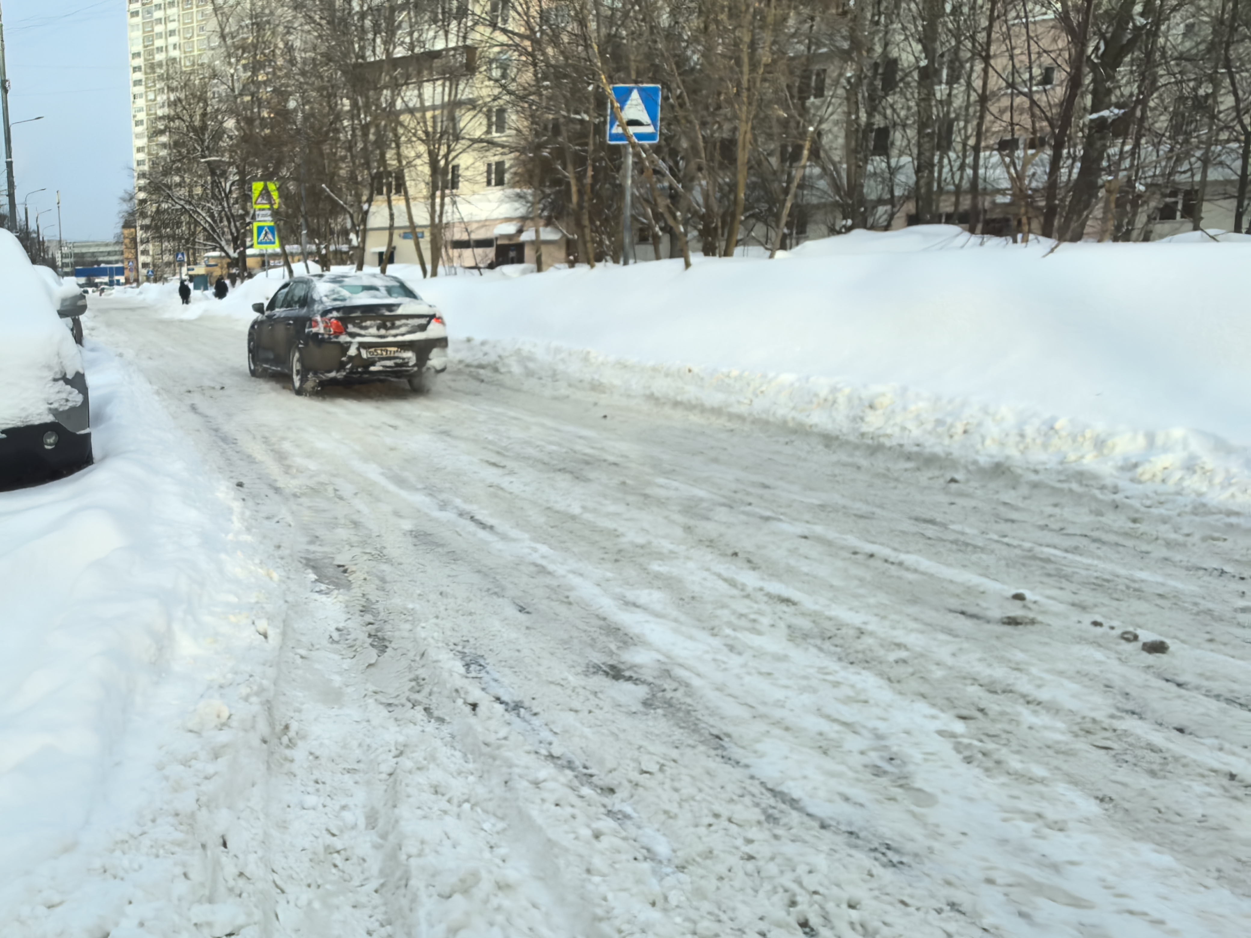 Проезжая часть без пешеходного перехода, покрытая снегом и льдом. На обочинах высокие сугробы, на дороге видны следы от шин. Видны знаки пешеходного перехода впереди, но сам переход не виден.