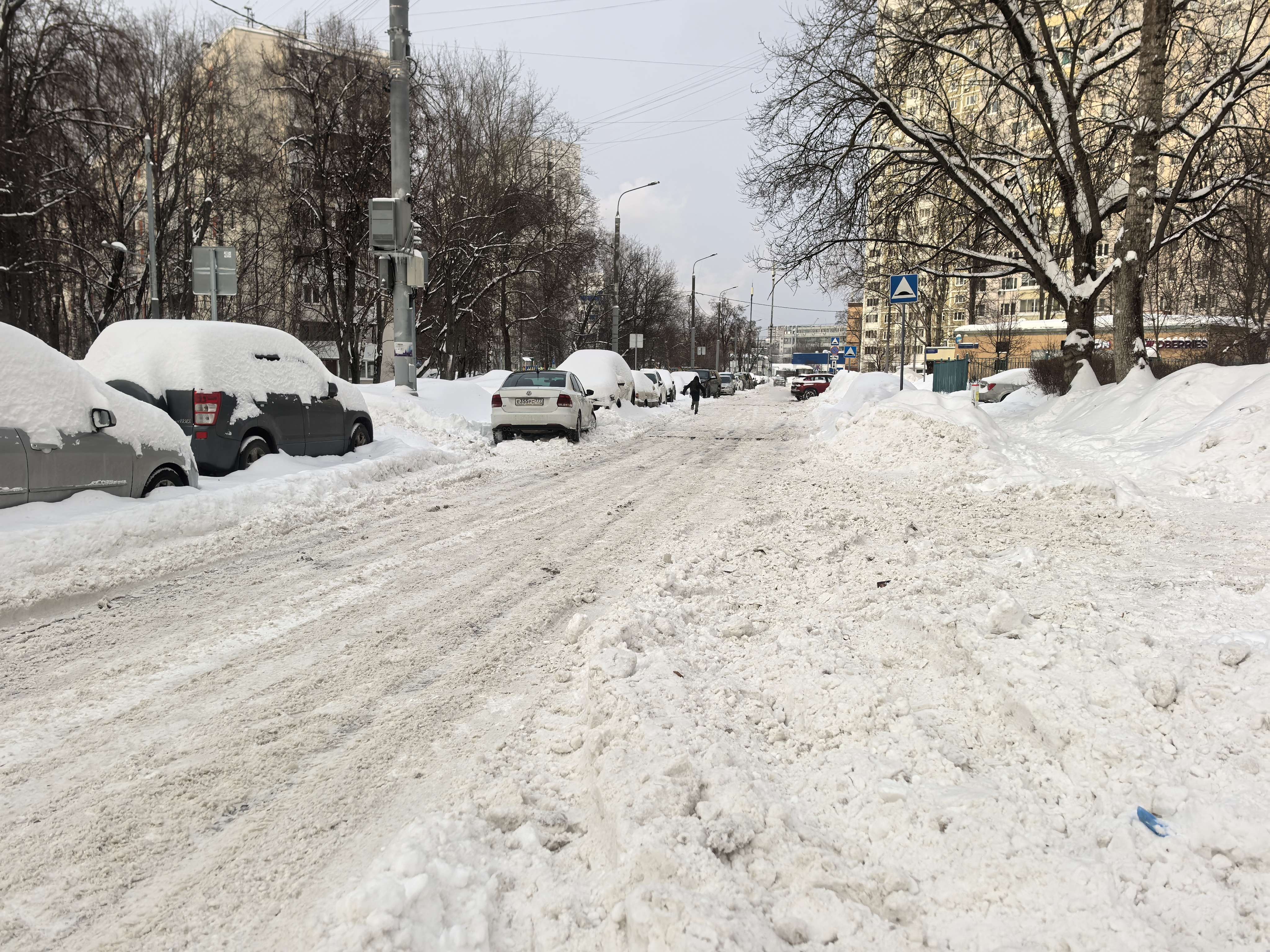 Городская улица в зимний период, покрытая снегом и наледью. Пешеходный переход отсутствует, тротуары полностью засыпаны снегом, что вынуждает пешеходов идти по проезжей части. Парковка автомобилей вдоль дороги блокирует обзор и сужает проезжую часть.