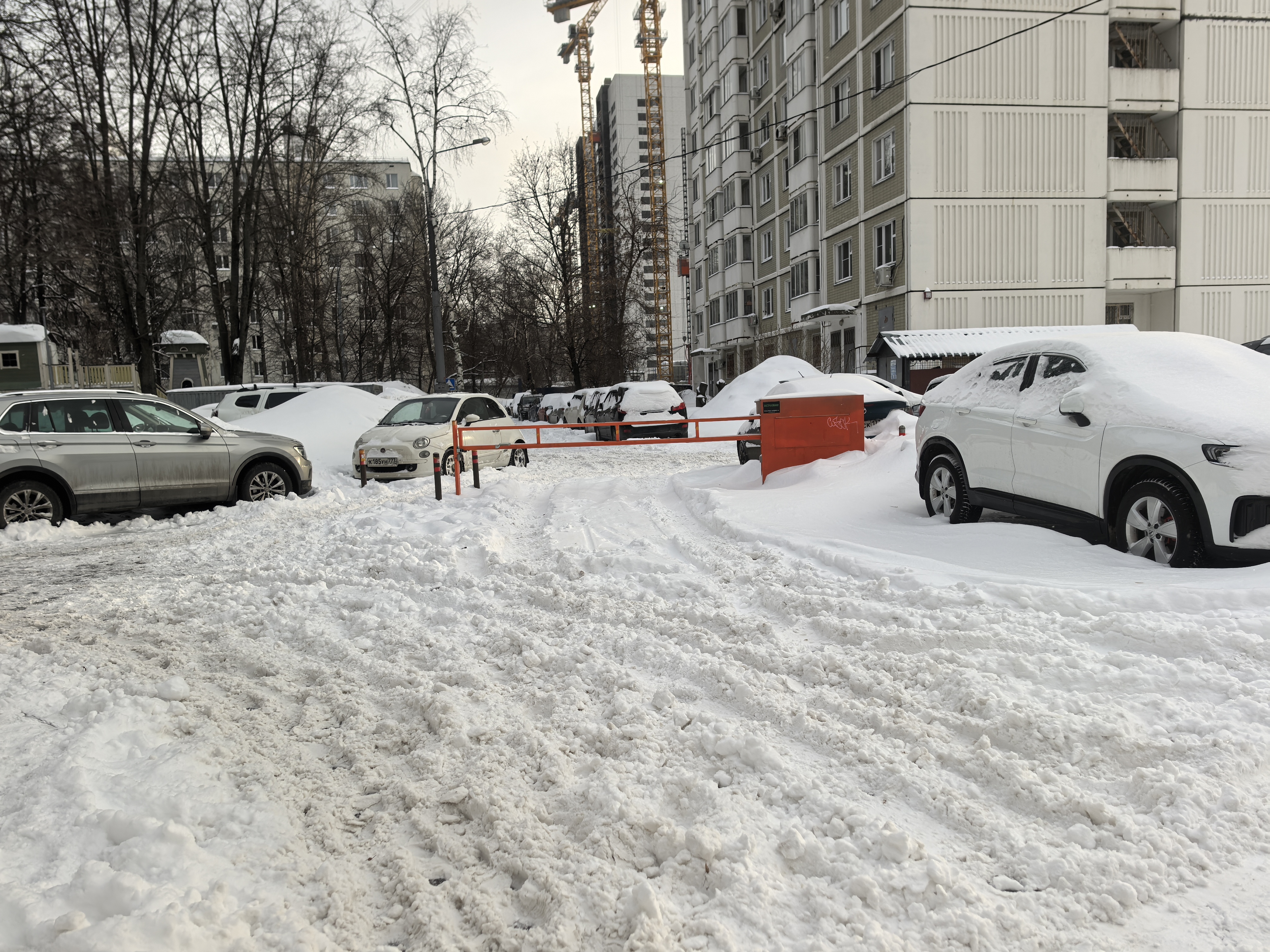 Дворовая территория, покрытая снегом, с припаркованными автомобилями и ограждением. Проезжая часть не просматривается из-за снежного покрова.