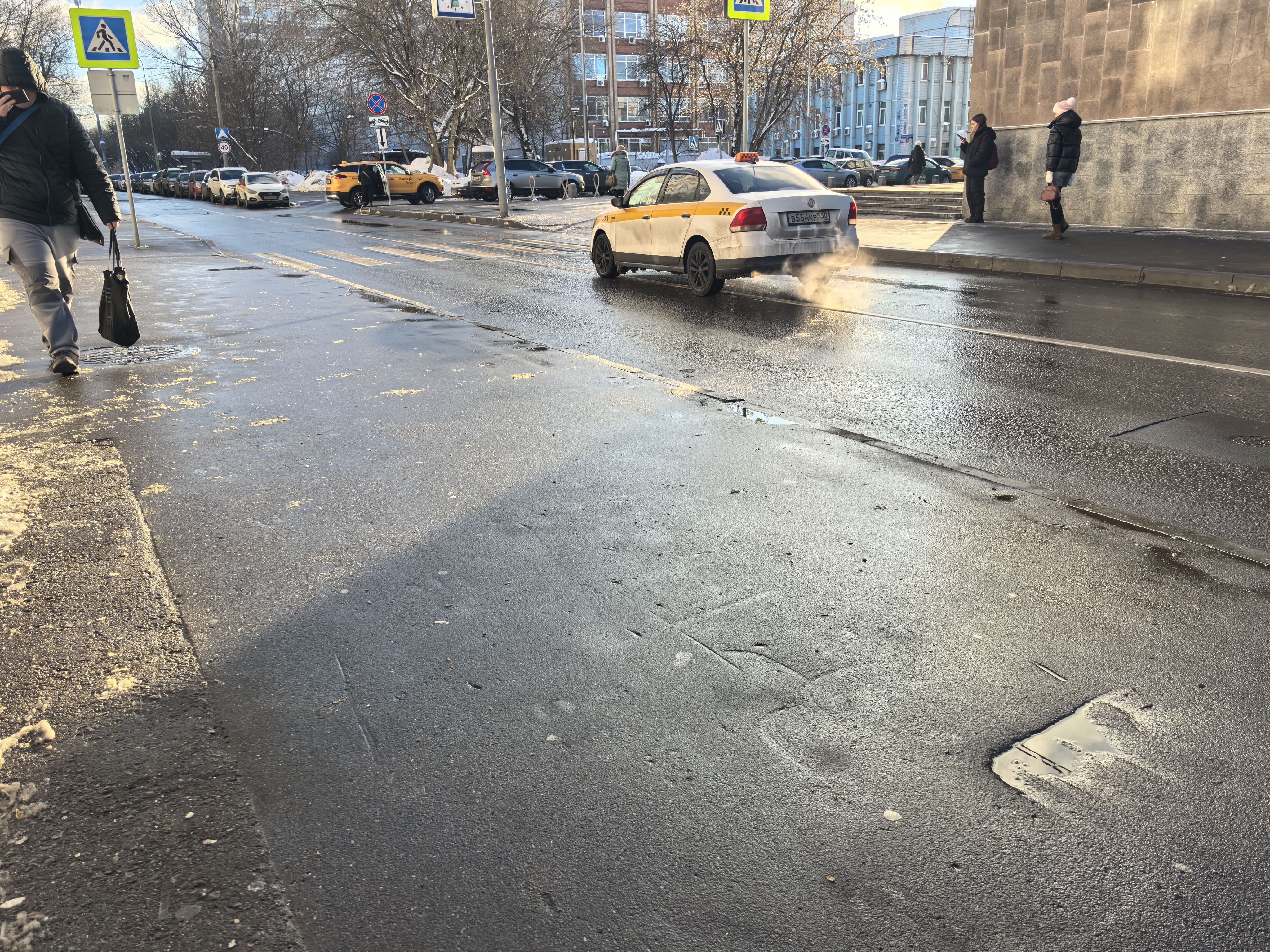 Городская улица с пешеходным переходом, обозначенным знаками и разметкой. Дорога асфальтированная, мокрая, с видимой разметкой.