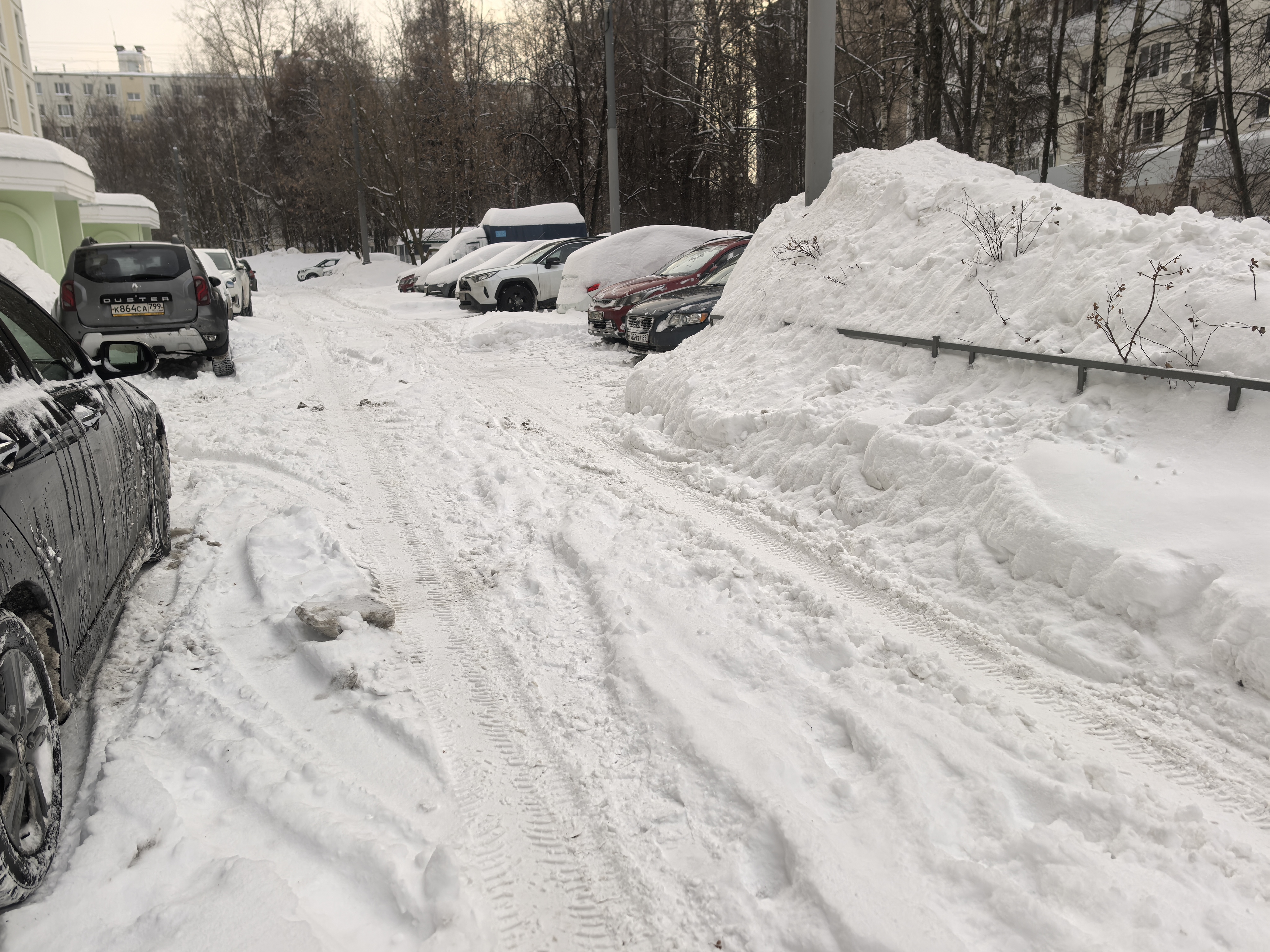 Зимняя дворовая территория с припаркованными автомобилями, проезжая часть полностью покрыта снегом и следами шин. Тротуары и обочины заблокированы высокими сугробами.