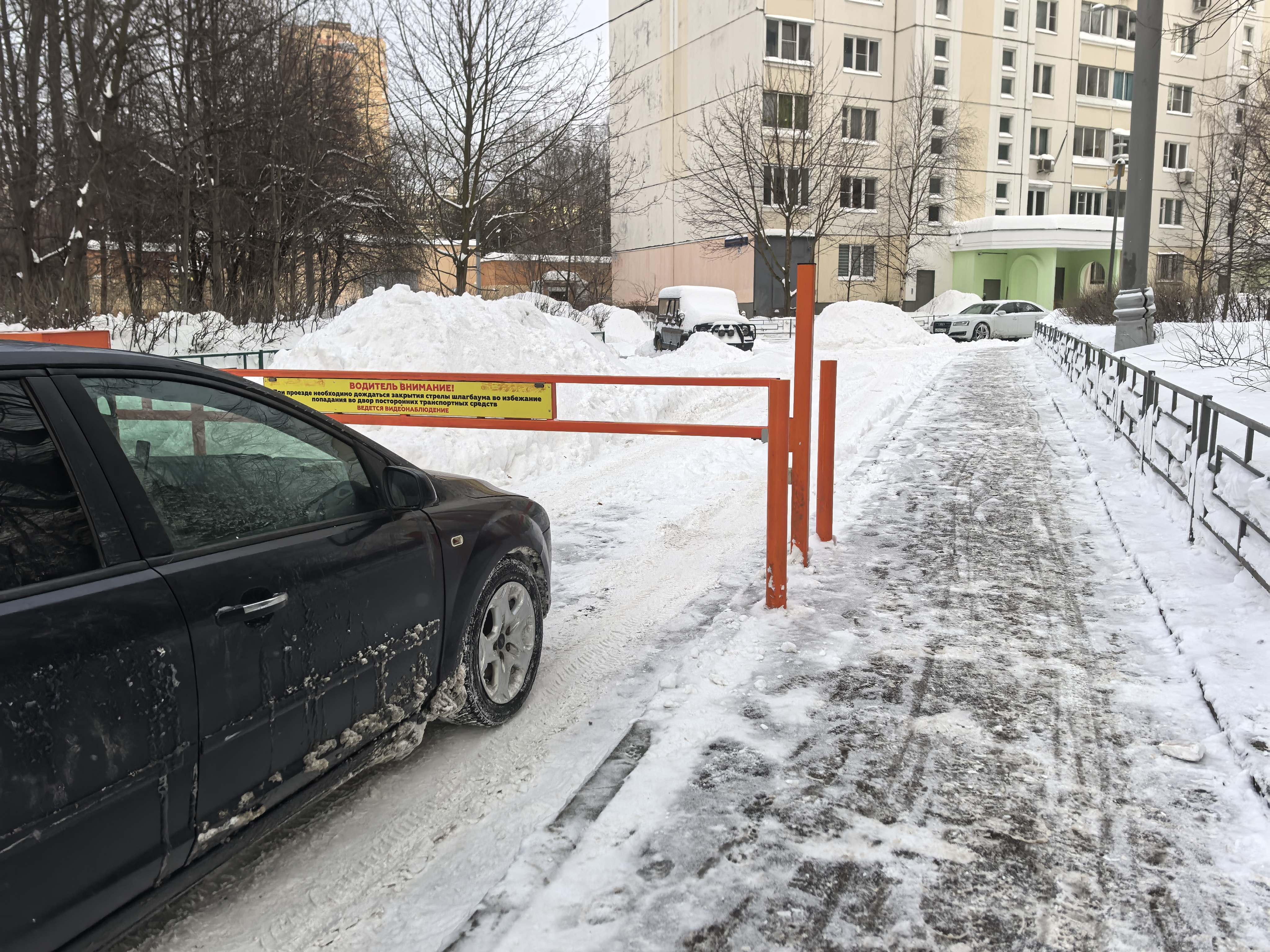 Дворовая территория с шлагбаумом, покрытая снегом и льдом. Видны припаркованные автомобили и жилой дом на заднем плане.
