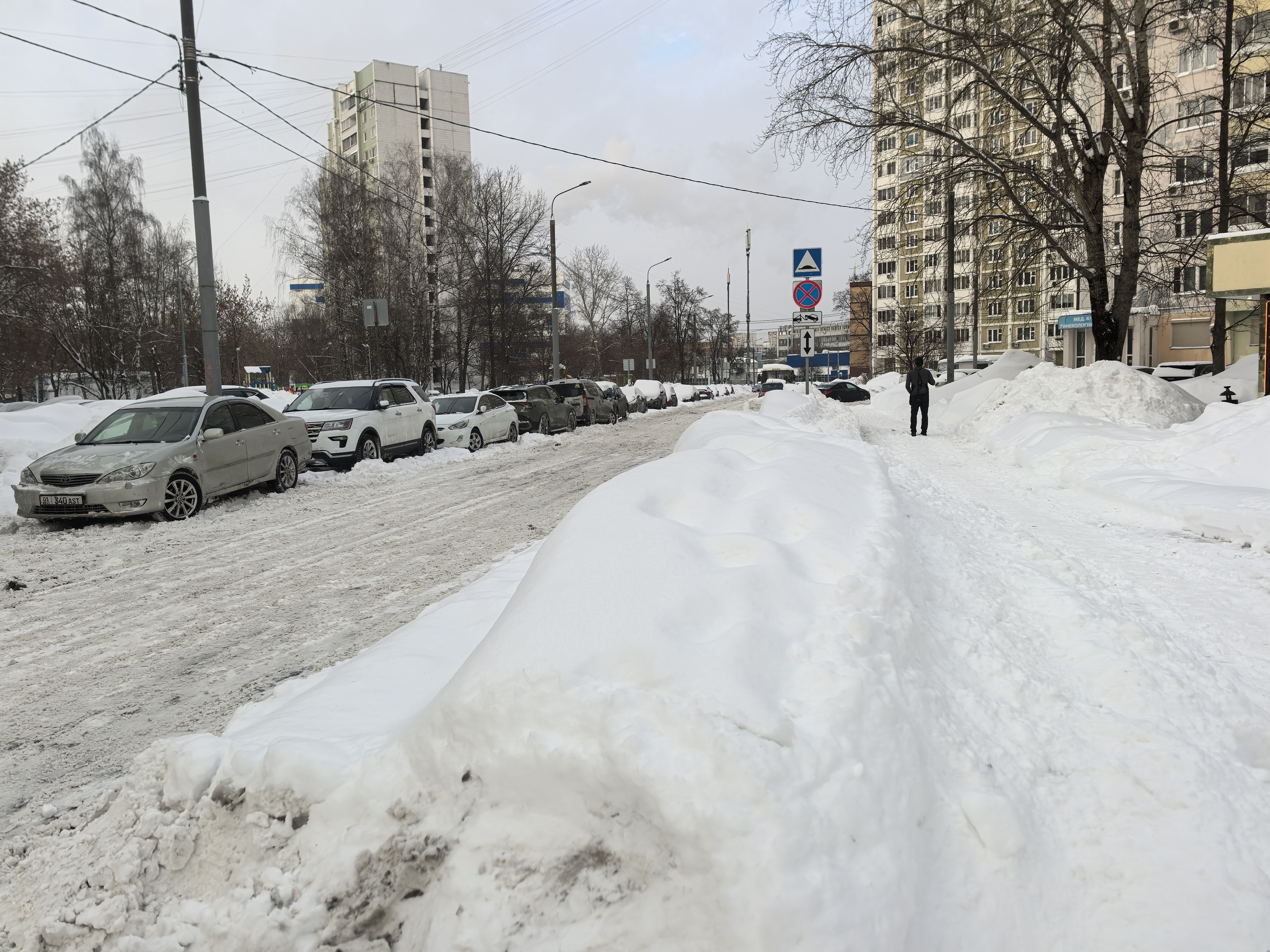 Участок городской улицы в зимний период. Видна проезжая часть, покрытая снегом и слякотью, с припаркованными автомобилями. Справа находится тротуар, частично засыпанный снегом, по которому идет пешеход. Вдали видны знаки, указывающие на приближение пешехо