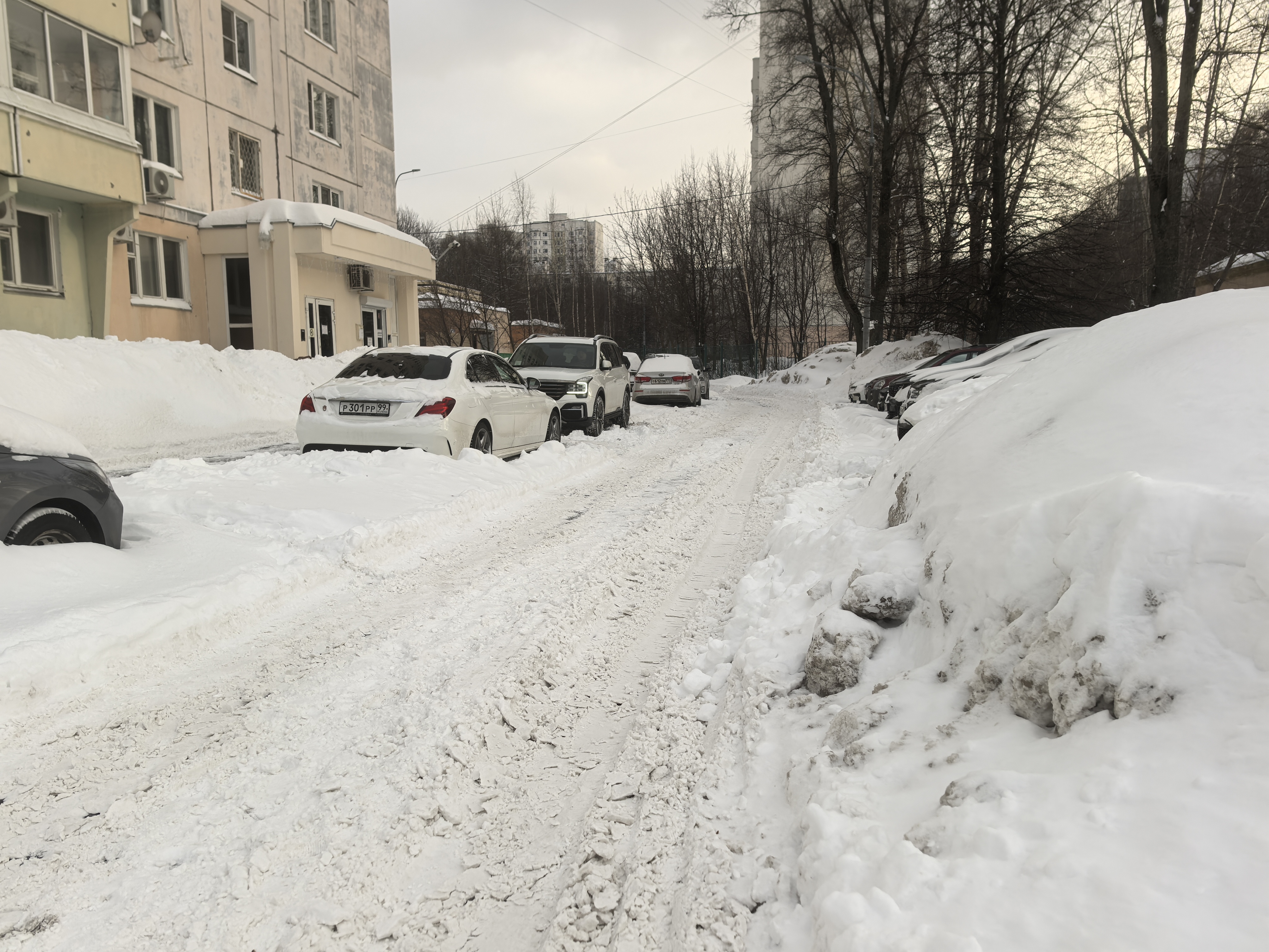 Дворовая территория, заснеженная проезжая часть с припаркованными автомобилями, тротуары полностью скрыты под снегом.