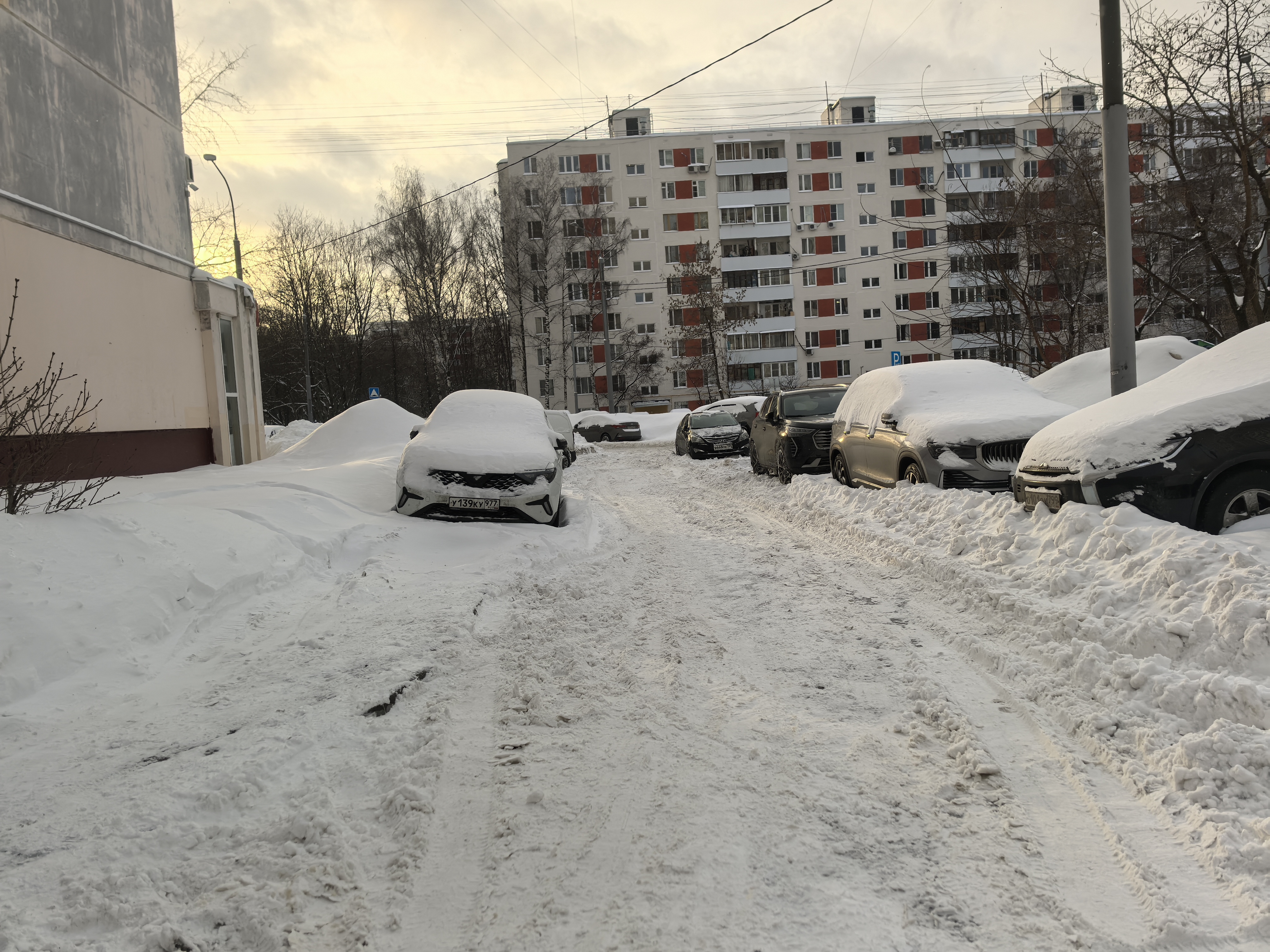 Дворовая территория с припаркованными автомобилями, покрытая снегом. Проезжая часть узкая, бордюры и тротуары не видны или полностью засыпаны снегом.