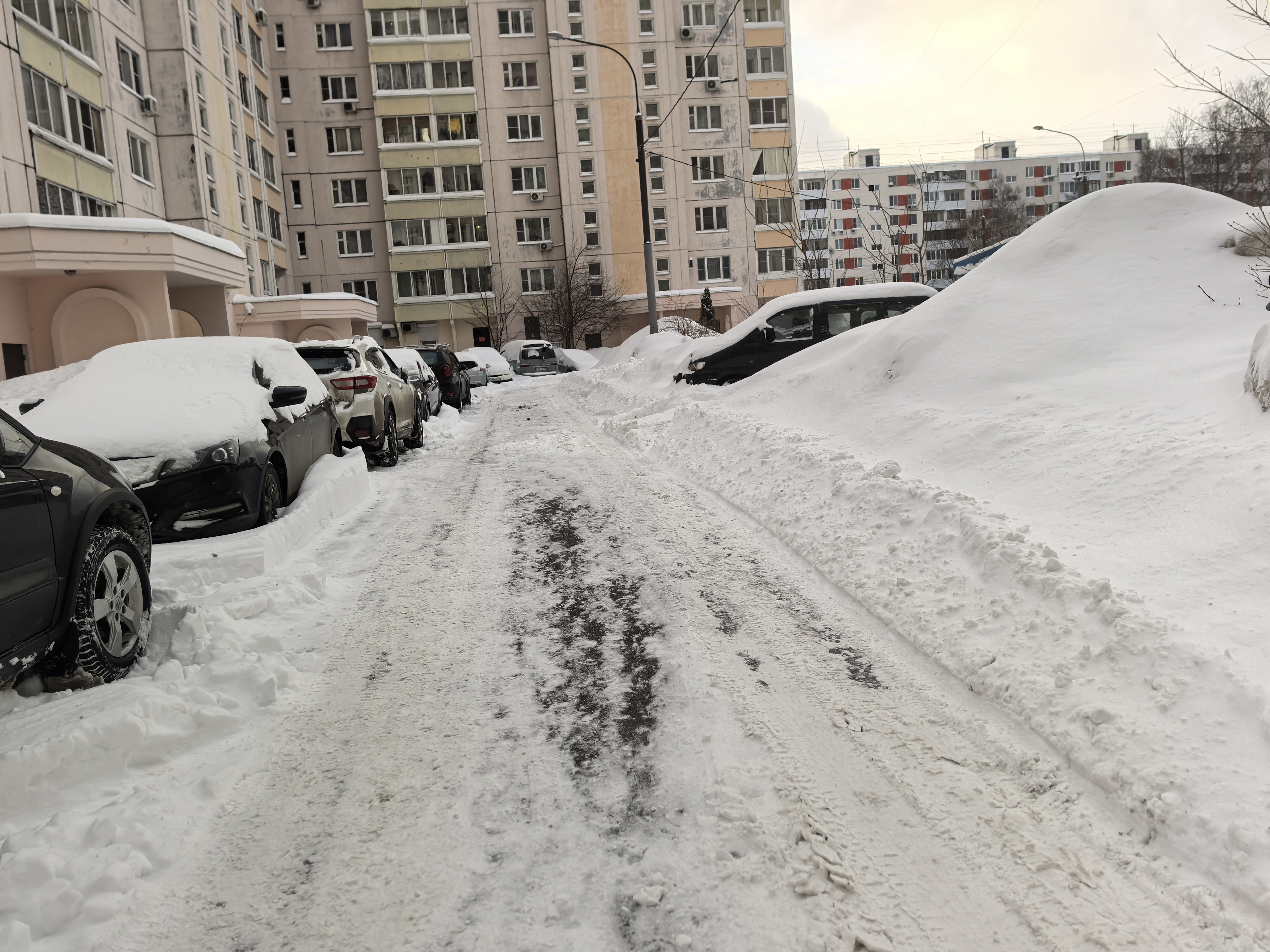 Дскальная территория между жилыми домами, покрытая снегом, с припаркованными автомобилями по обеим сторонам проезжей части.