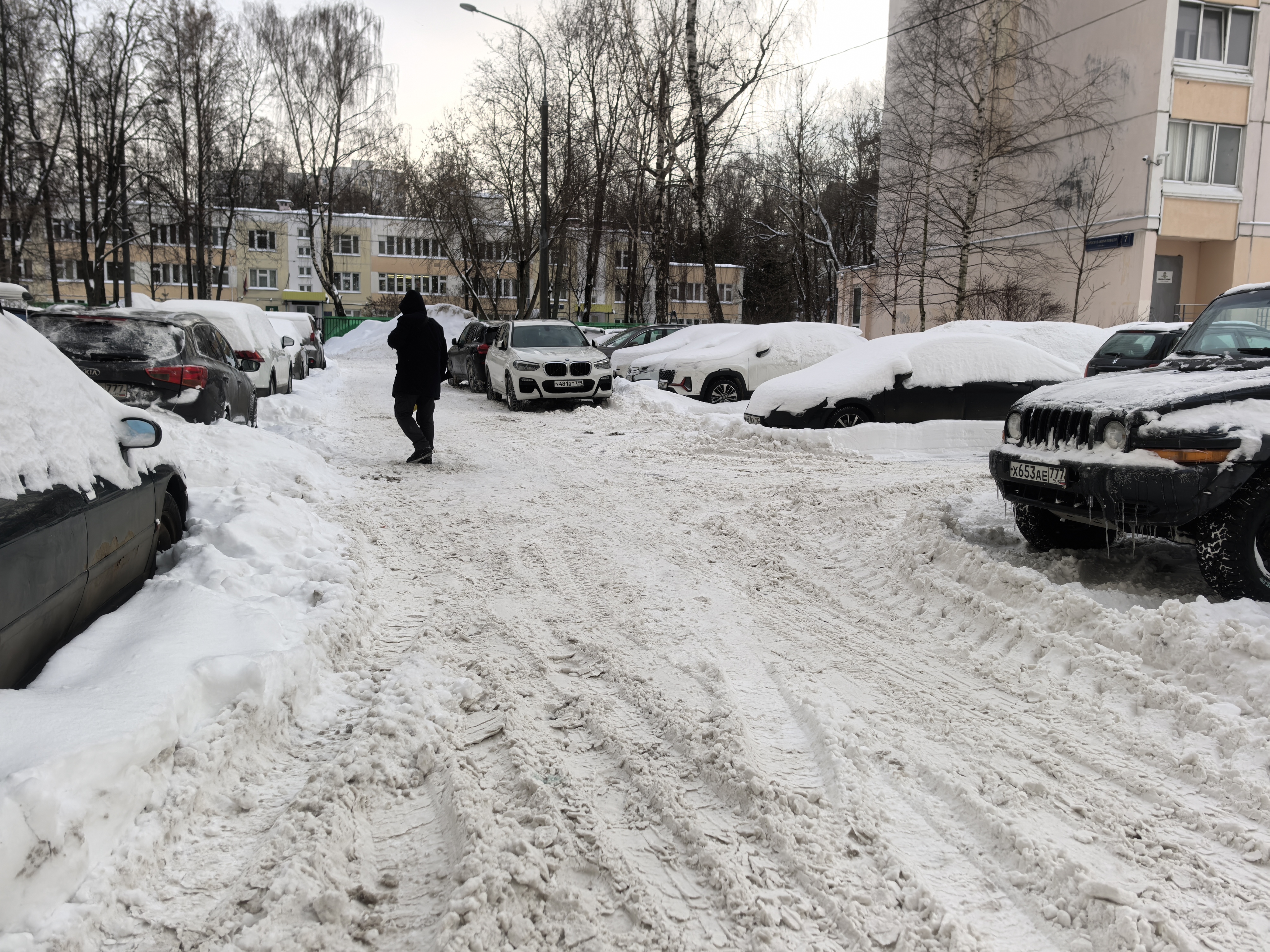 Дворовая территория между жилыми домами, покрытая снегом, с припаркованными автомобилями по обеим сторонам проезжей части.