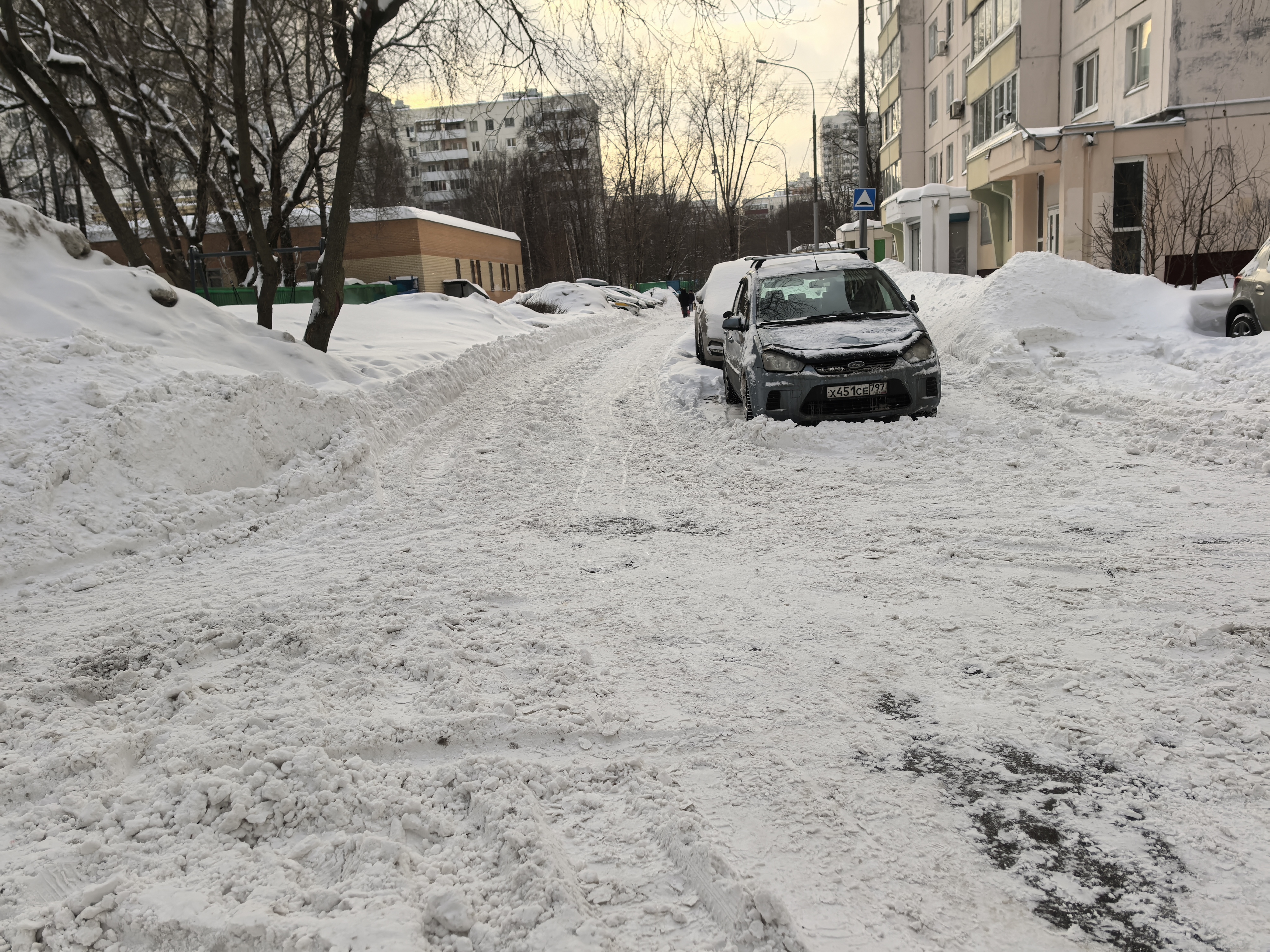 Дворовая территория, покрытая снегом, с припаркованными автомобилями вдоль проезжей части. Пешеходных переходов и бордюров не видно.