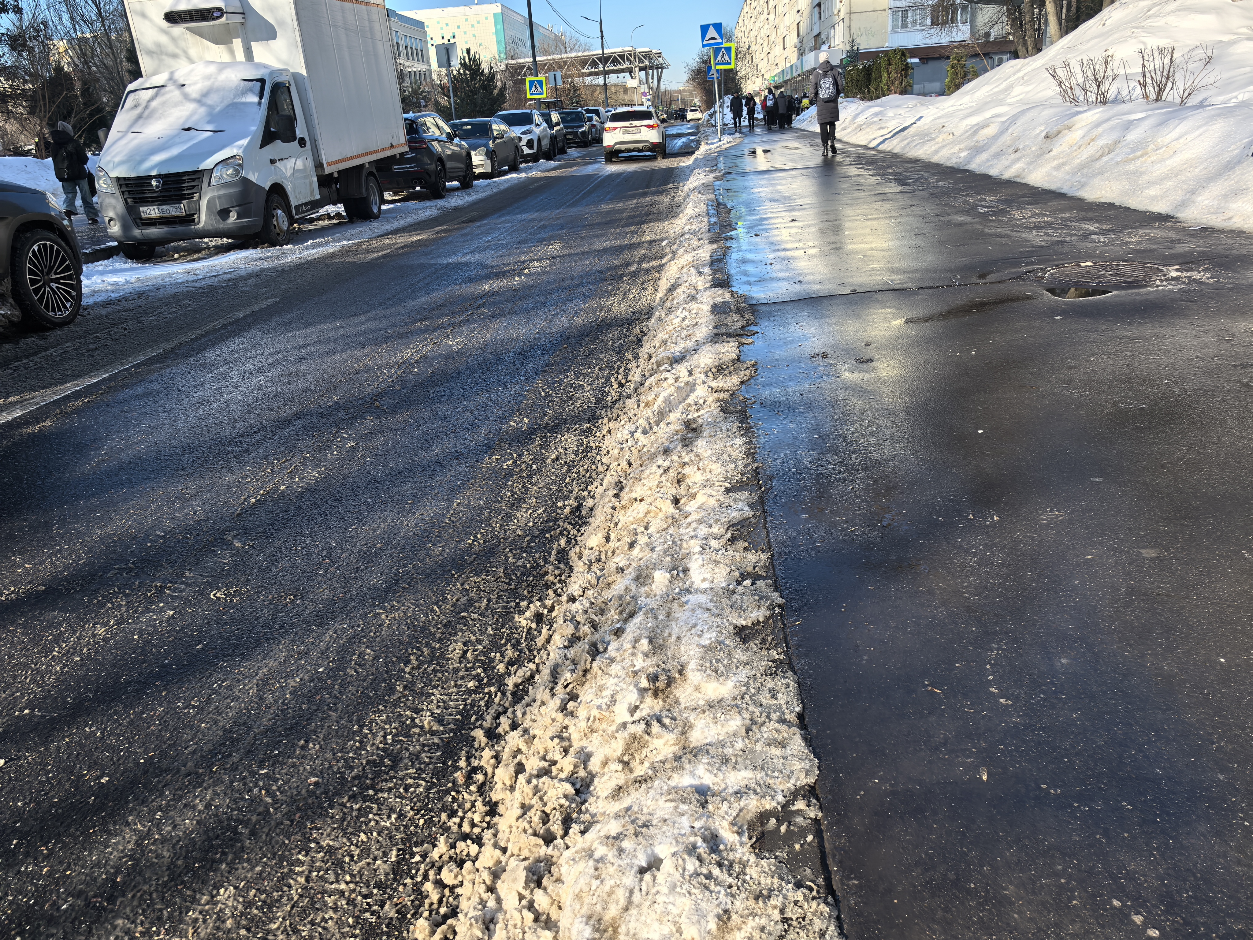 Участок городской дороги с пешеходным переходом, тротуаром и припаркованными автомобилями. Видны пешеходы и дорожные знаки.