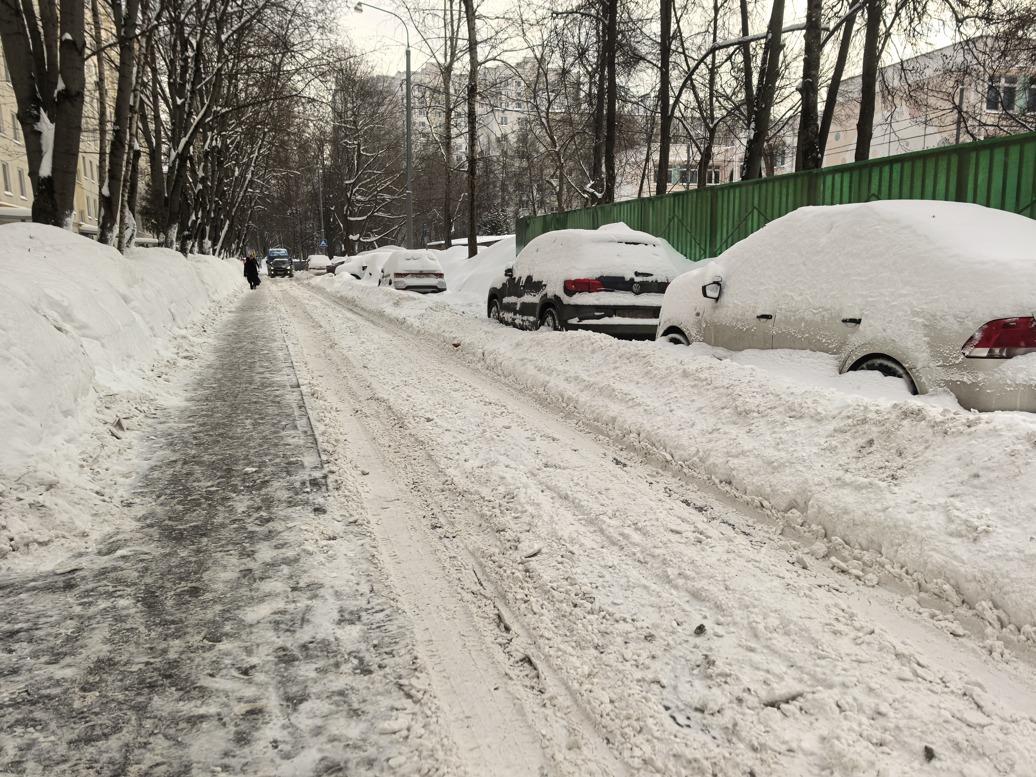 Проезжая часть без организованного пешеходного перехода, покрытая снегом и льдом, с припаркованными автомобилями, частично блокирующими обзор.