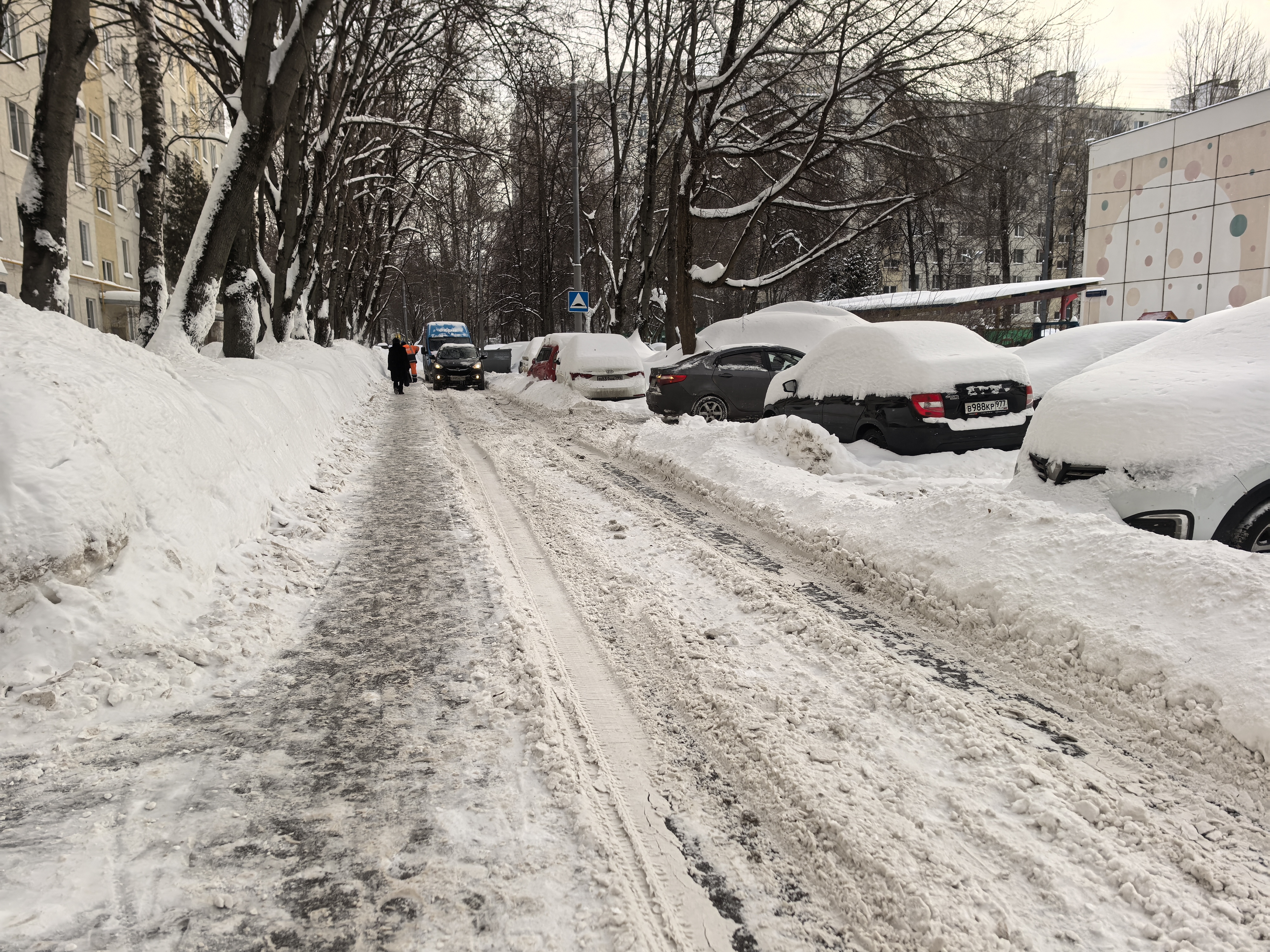 Участок проезжей части в зимний период, покрытый снегом и льдом. Тротуары засыпаны снегом, припаркованные автомобили также покрыты снегом. Видна пешеходная дорожка, вытопленная в снегу, но она находится в непосредственной близости от проезжей части. На за