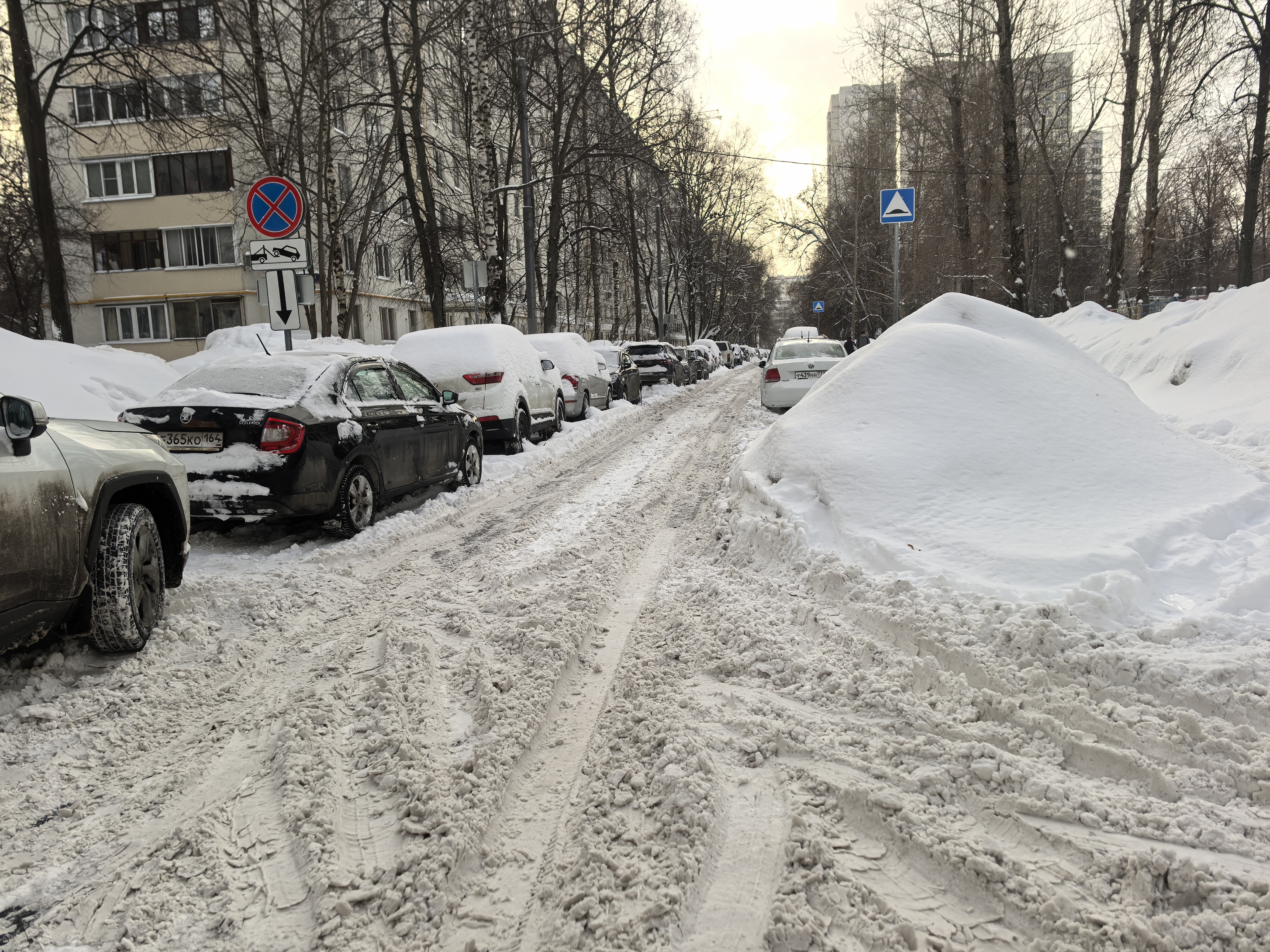 Участок проезжей части в жилом районе, покрытый снегом и ледяной кашей. Пешеходный переход отсутствует. Тротуары полностью засыпаны снегом, бордюры не видны.