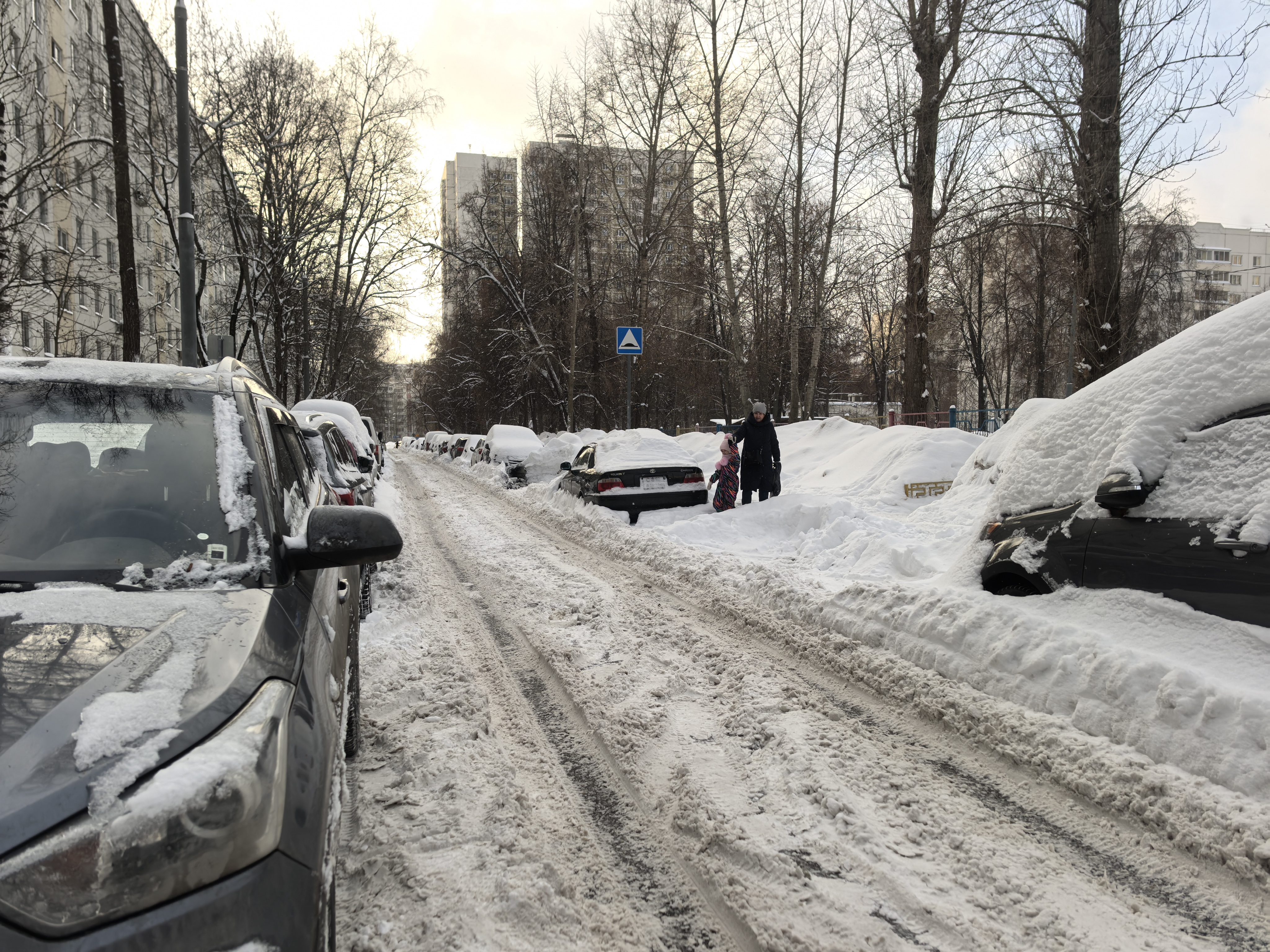 Участок дороги во дворе или на прилегающей территории, покрытый снегом. Видны припаркованные автомобили, тротуары засыпаны снегом, пешеходы идут по снегу. Пешеходного перехода (зебры) на видимом участке нет, но есть знак «Пешеходный переход» впереди.
