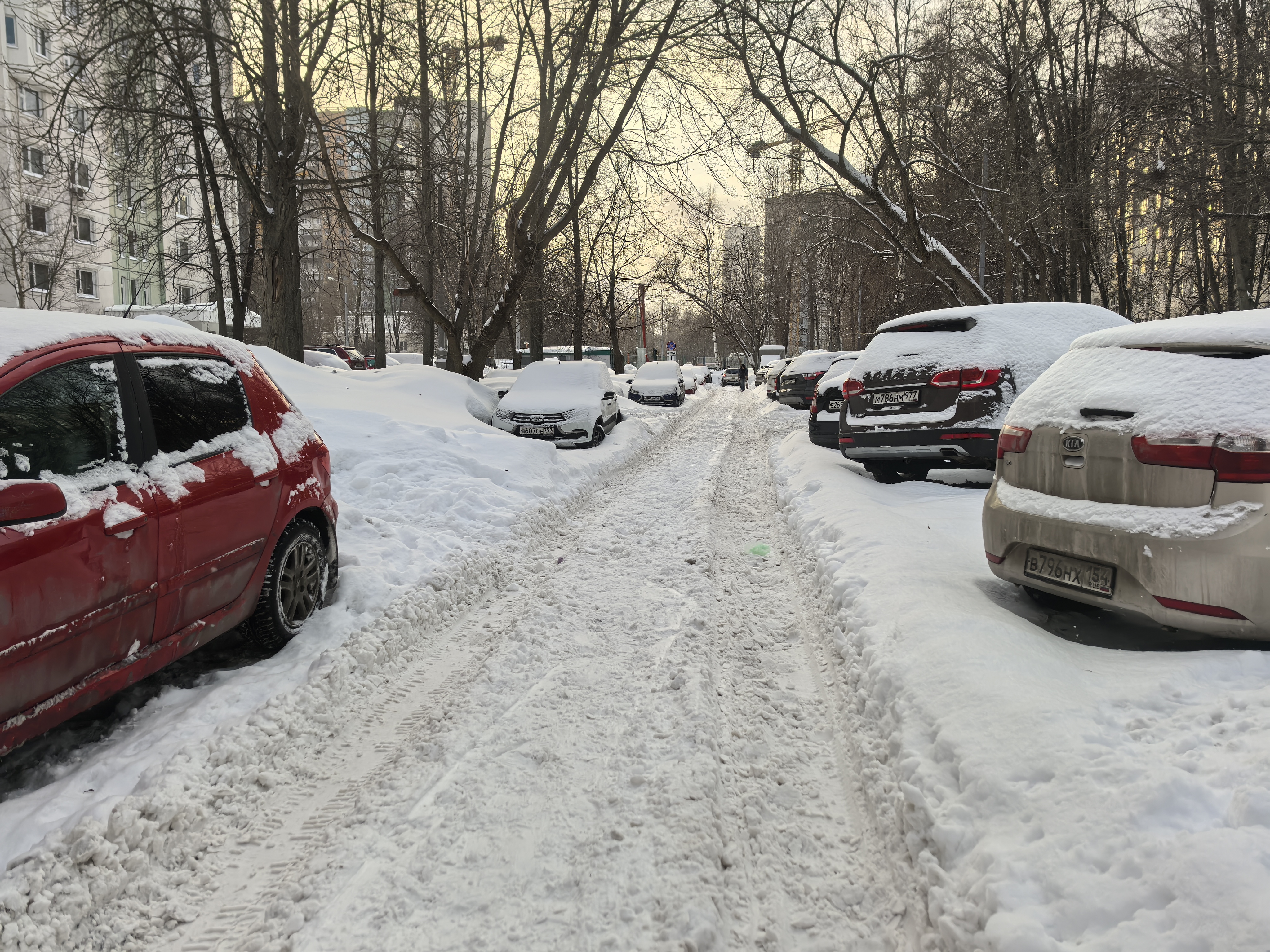 Дворовая территория с парковкой автомобилей, покрытая снегом. Проезжая часть узкая, тротуары полностью засыпаны снегом.