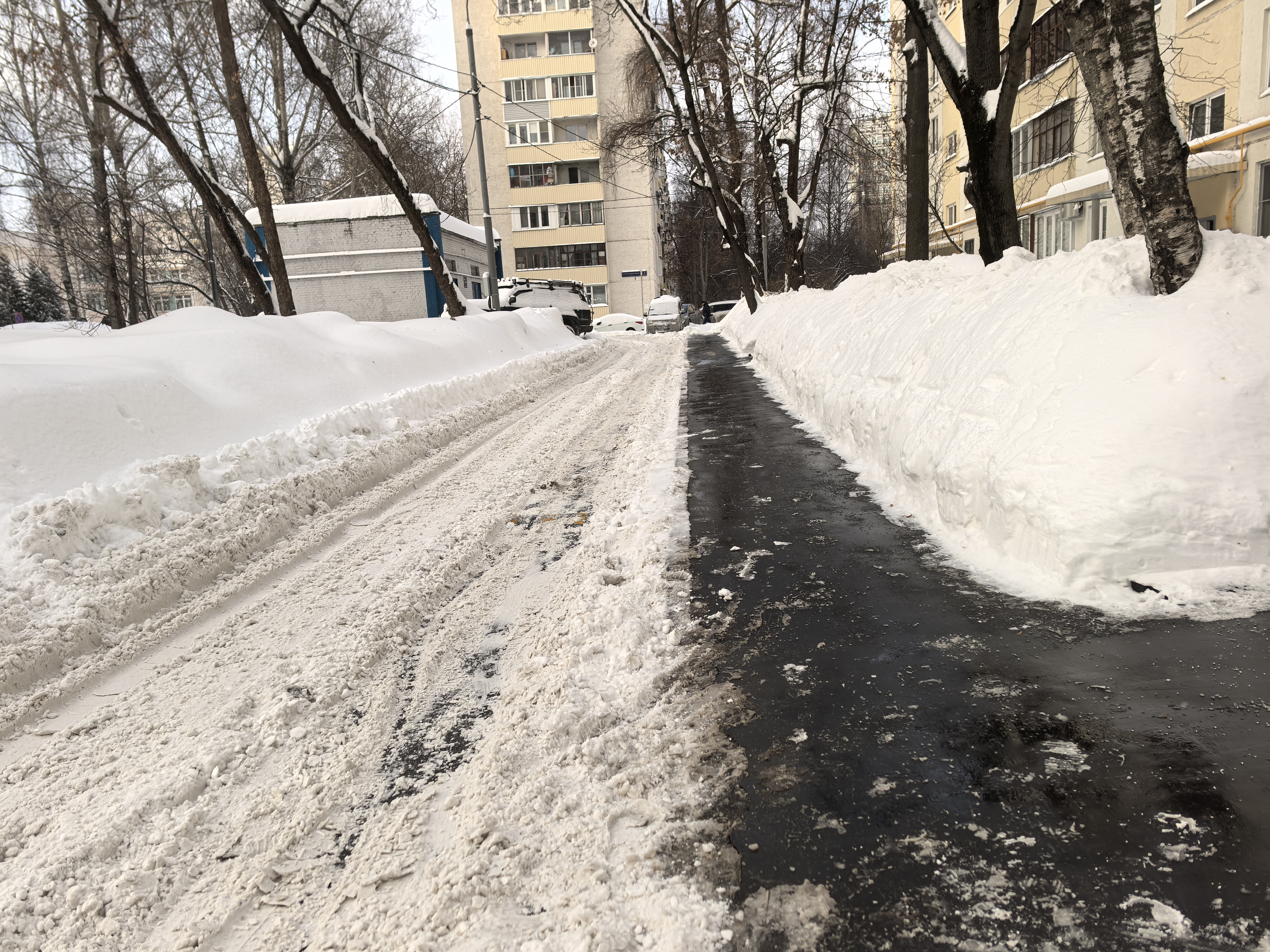 Участок дороги во дворе жилого массива с заснеженной проезжей частью и узкой пешеходной дорожкой, очищенной от снега. Переход отсутствует.