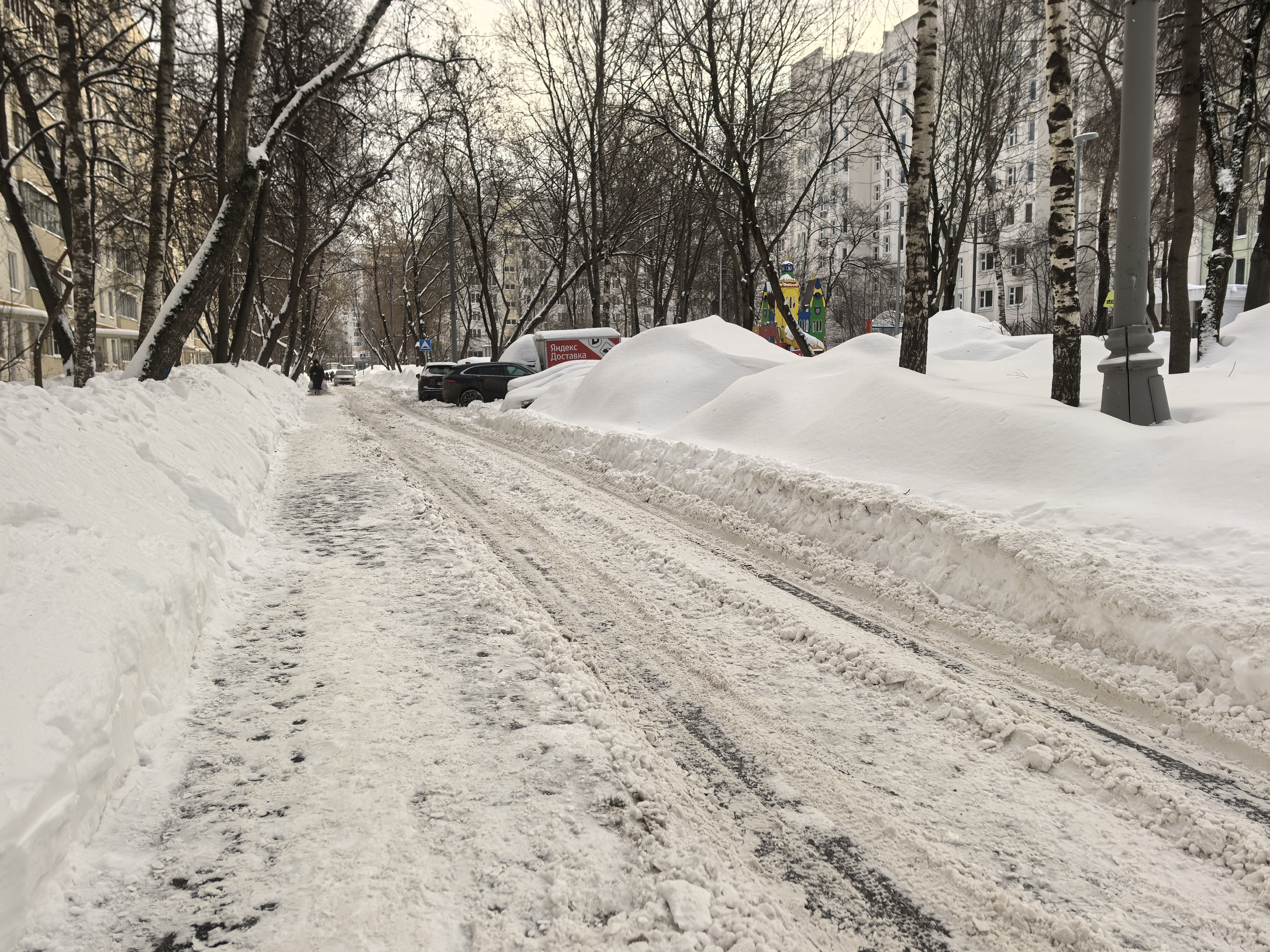 Дворовая территория, покрытая снегом, с припаркованными автомобилями и детской площадкой на заднем плане.