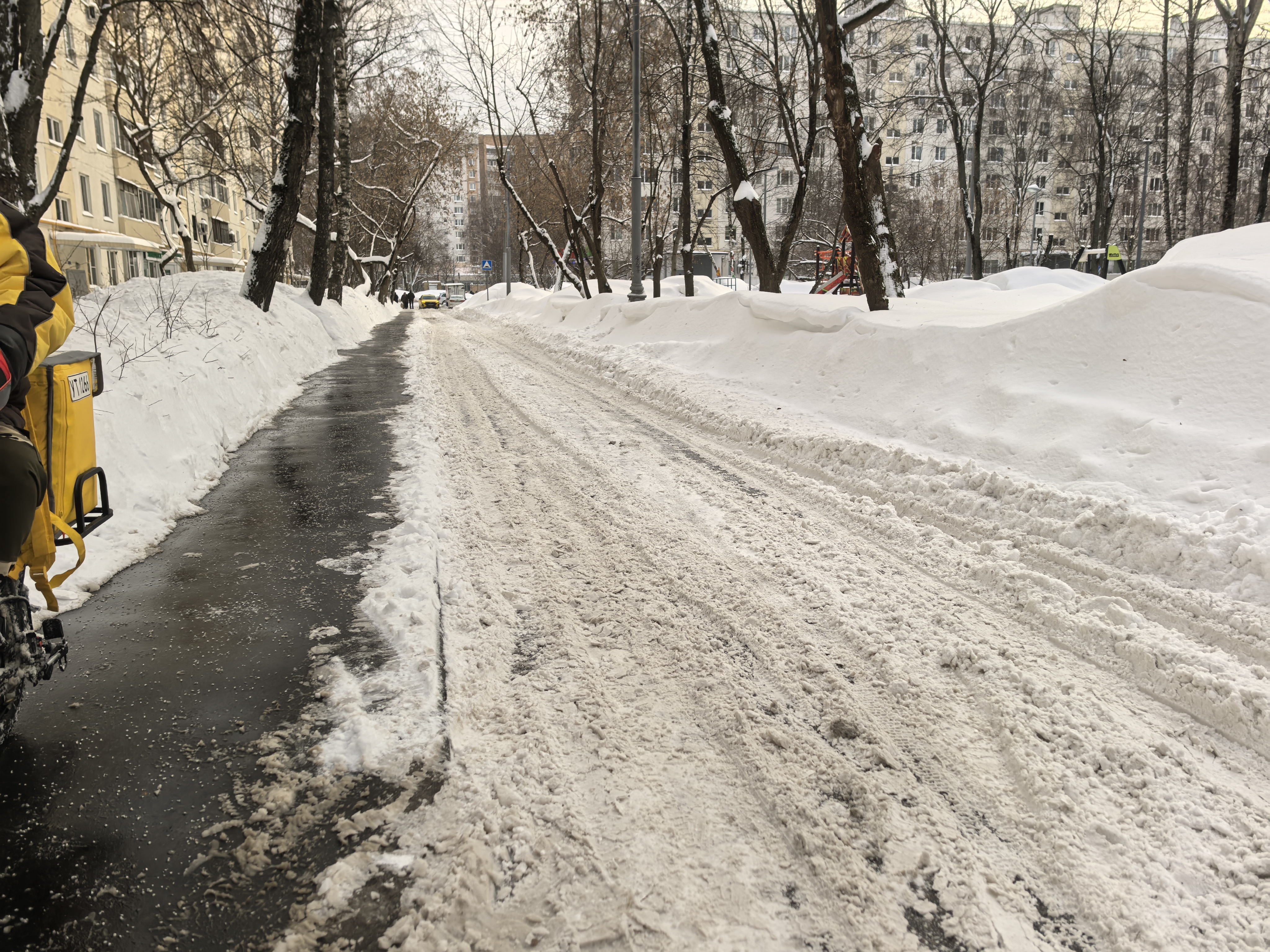 Участок дороги с узким пешеходным тротуаром, очищенным от снега, и широкой проезжей частью, покрытой снегом и колеей. Пешеходный переход виден вдалеке, но не является обустроенным на данном участке.