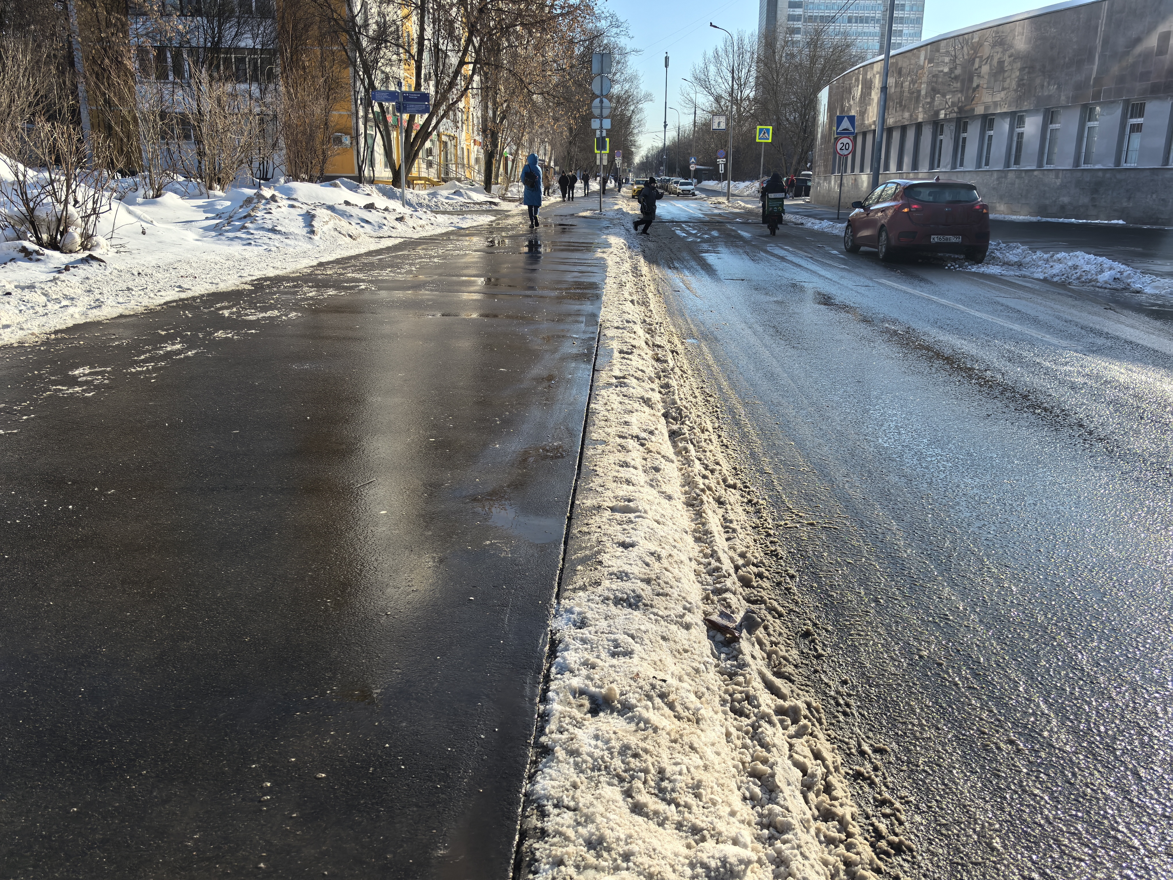 Участок городской улицы без пешеходного перехода. Видны тротуар, проезжая часть, разделенная снежной насыпью, и дорожные знаки. На тротуаре идут пешеходы, по дороге движется транспорт.
