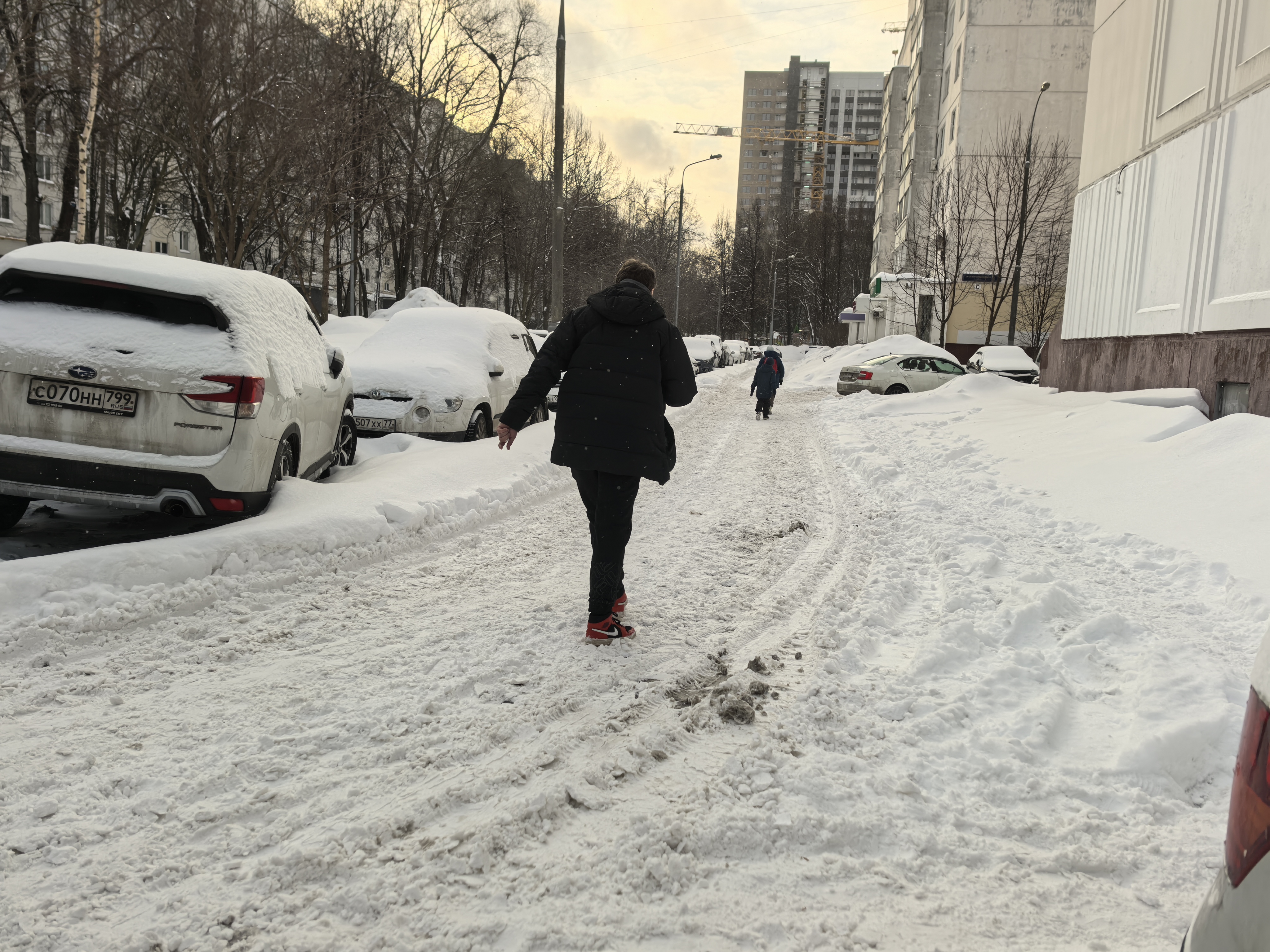 Дворовая территория, заснеженная проезжая часть между жилыми домами и припаркованными автомобилями. Пешеходы идут по снегу.