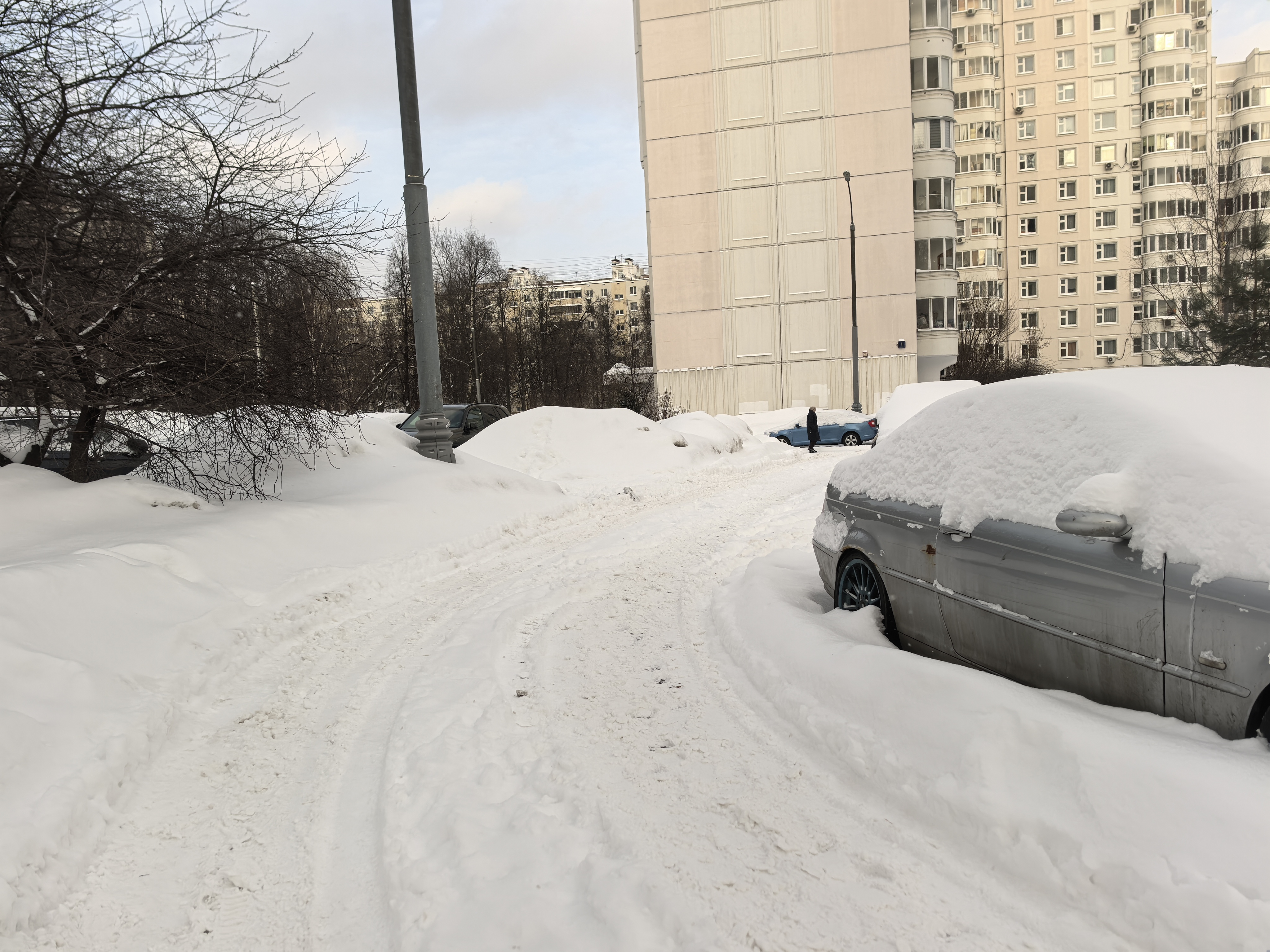 Дворовая территория, покрытая снегом, с припаркованными автомобилями и жилыми домами на заднем плане.