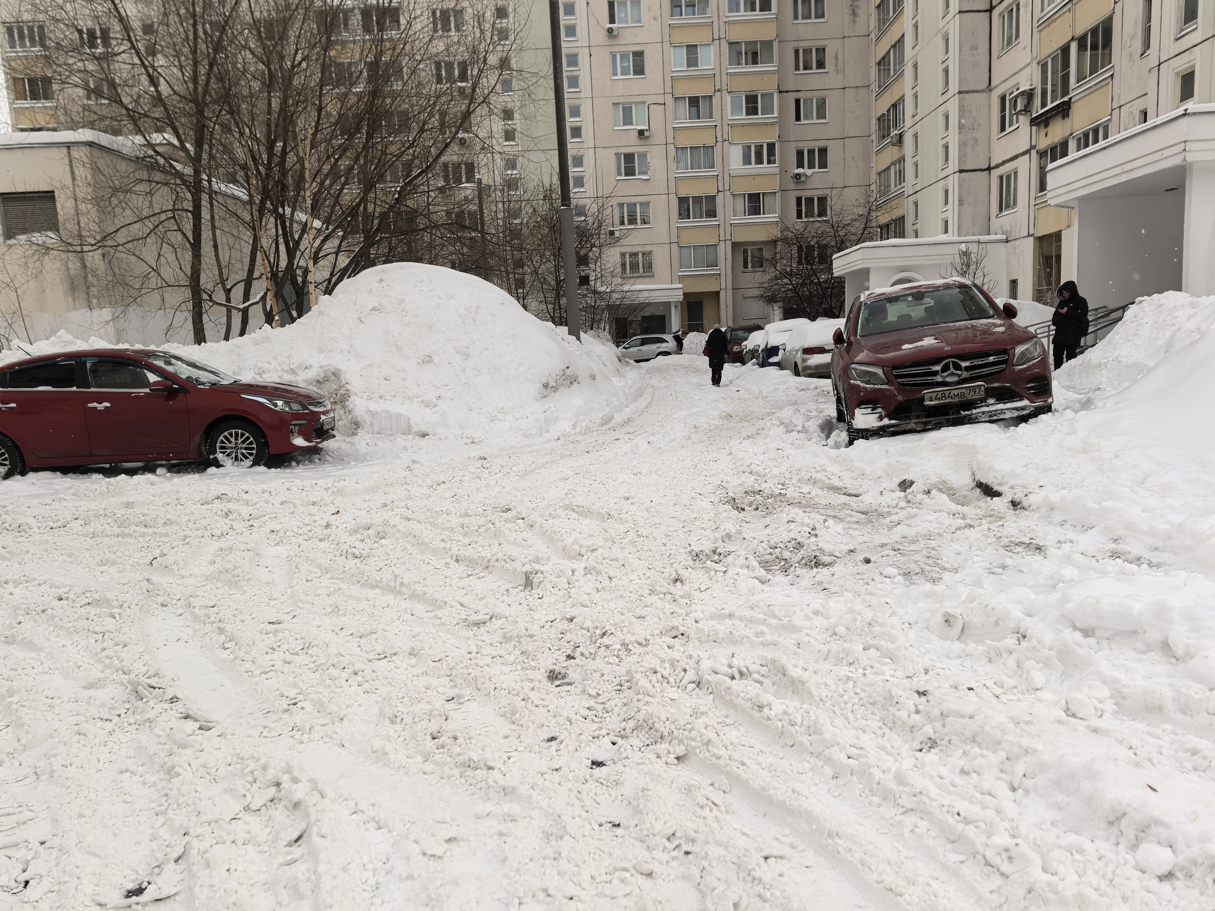 Дворовая территория, заснеженная дорога между жилыми домами. Видны припаркованные автомобили, один из которых (мерседес) частично заблокирован сугробом. Дорога покрыта снегом, разметка отсутствует.