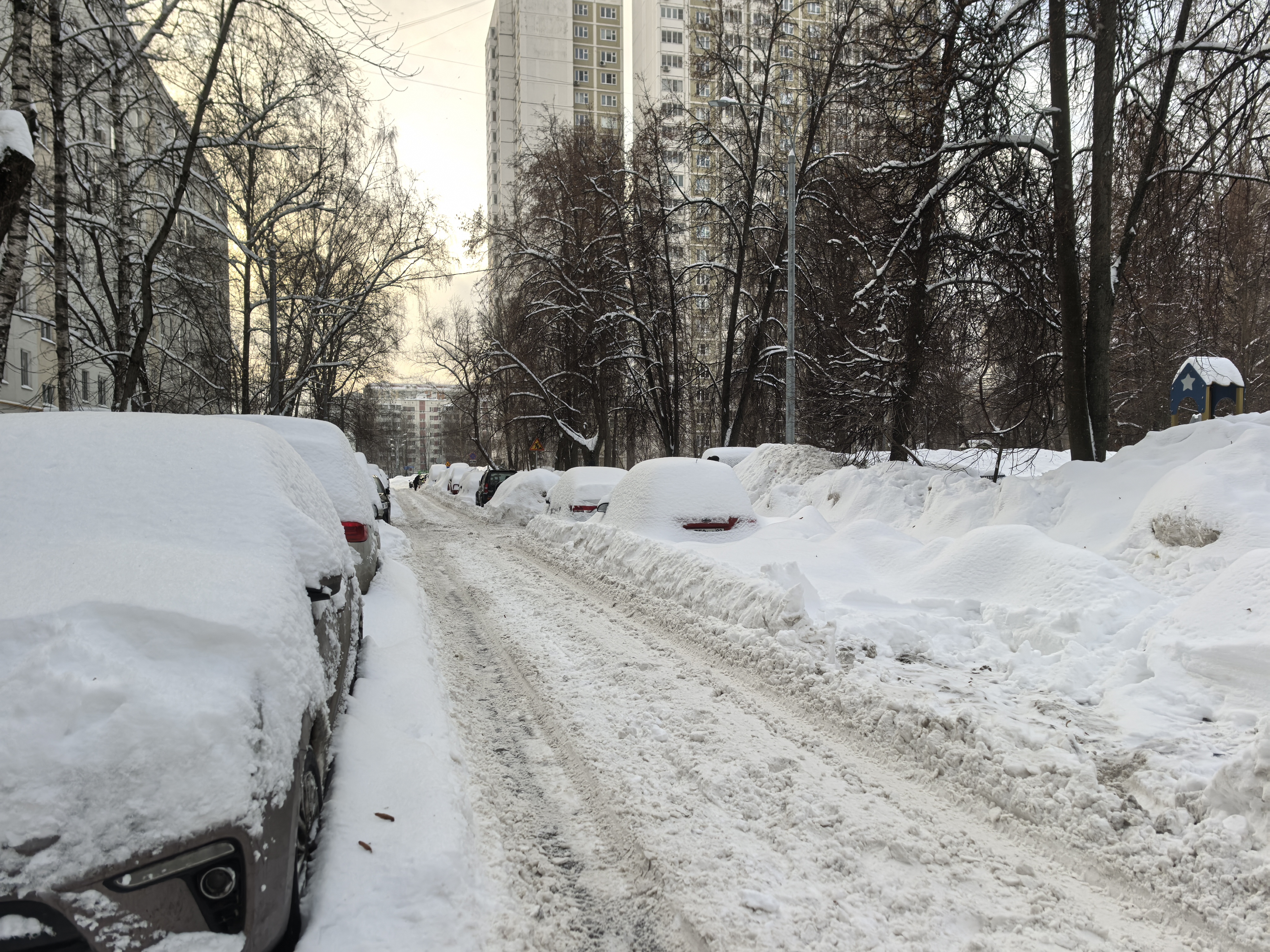 Дворовая территория с припаркованными автомобилями, покрытая снегом. Видны жилые дома и детская площадка справа.