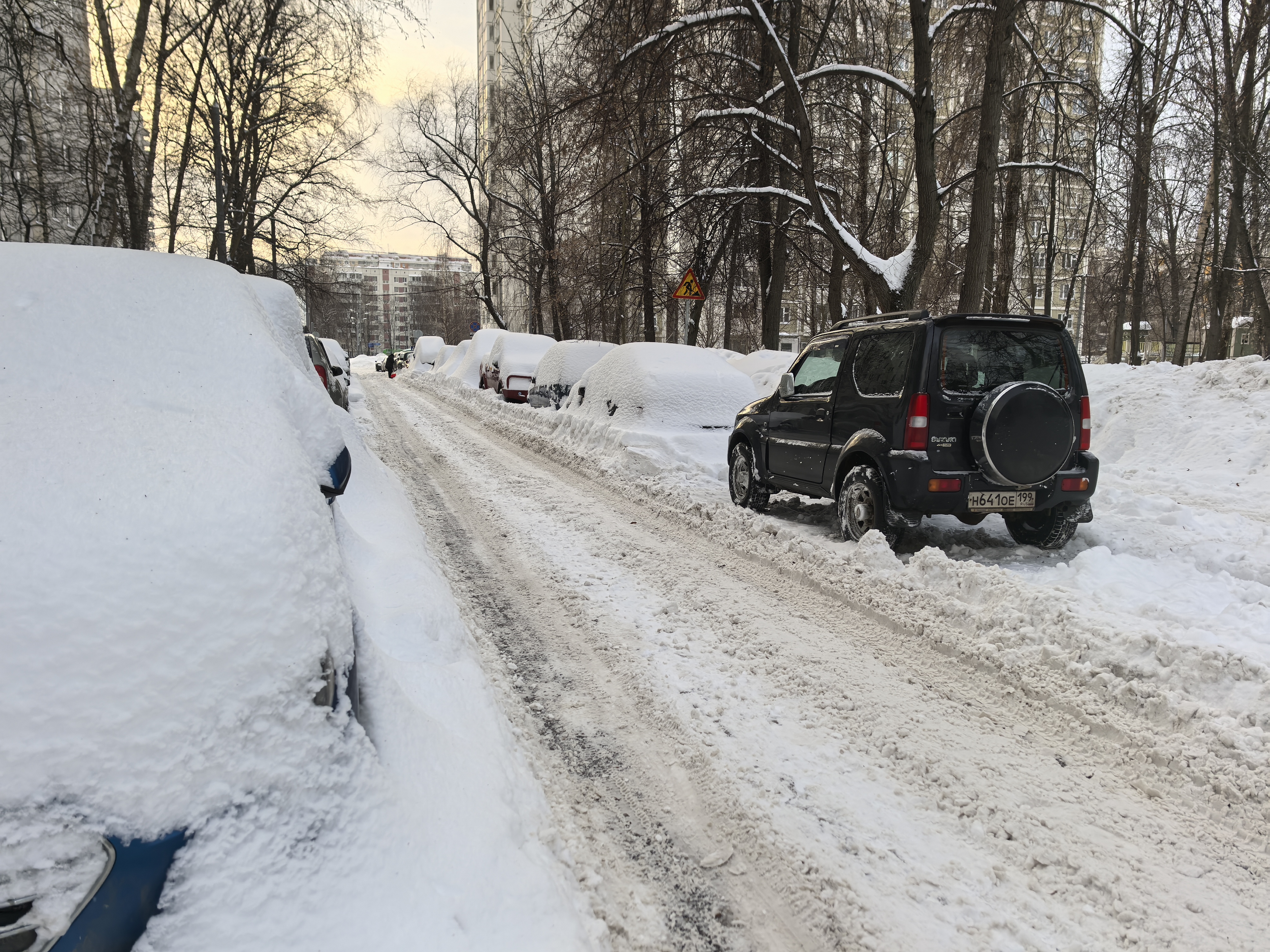 Участок дороги без пешеходного перехода, покрытый снегом и льдом. С обеих сторон припаркованы автомобили, полностью засыпанные снегом. На заднем плане виден знак «Дети».