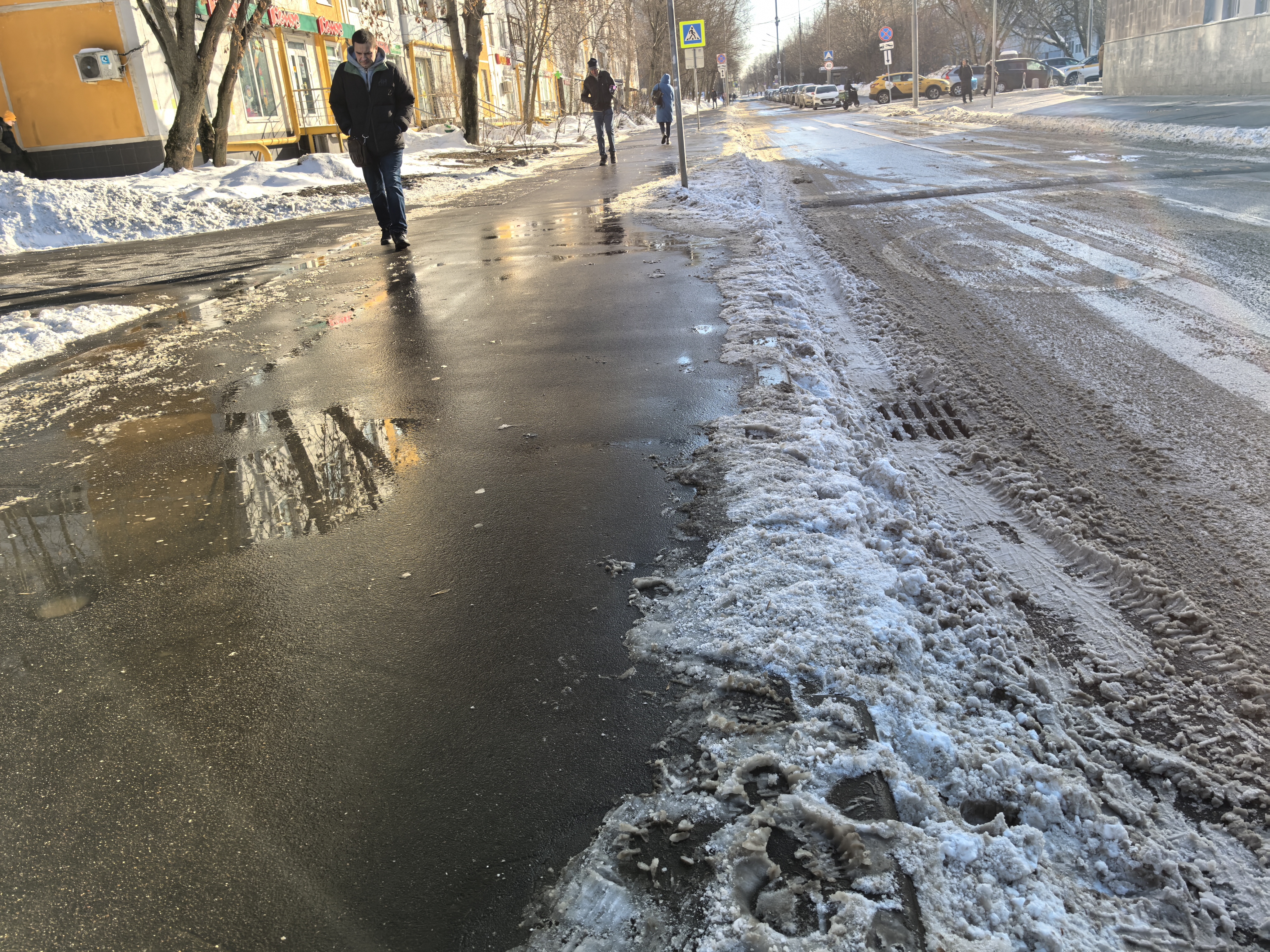 Городская улица с пешеходным переходом, тротуаром и прилегающими зданиями. Видны следы зимы: снег на обочинах, ледяная корка и лужи на проезжей части.