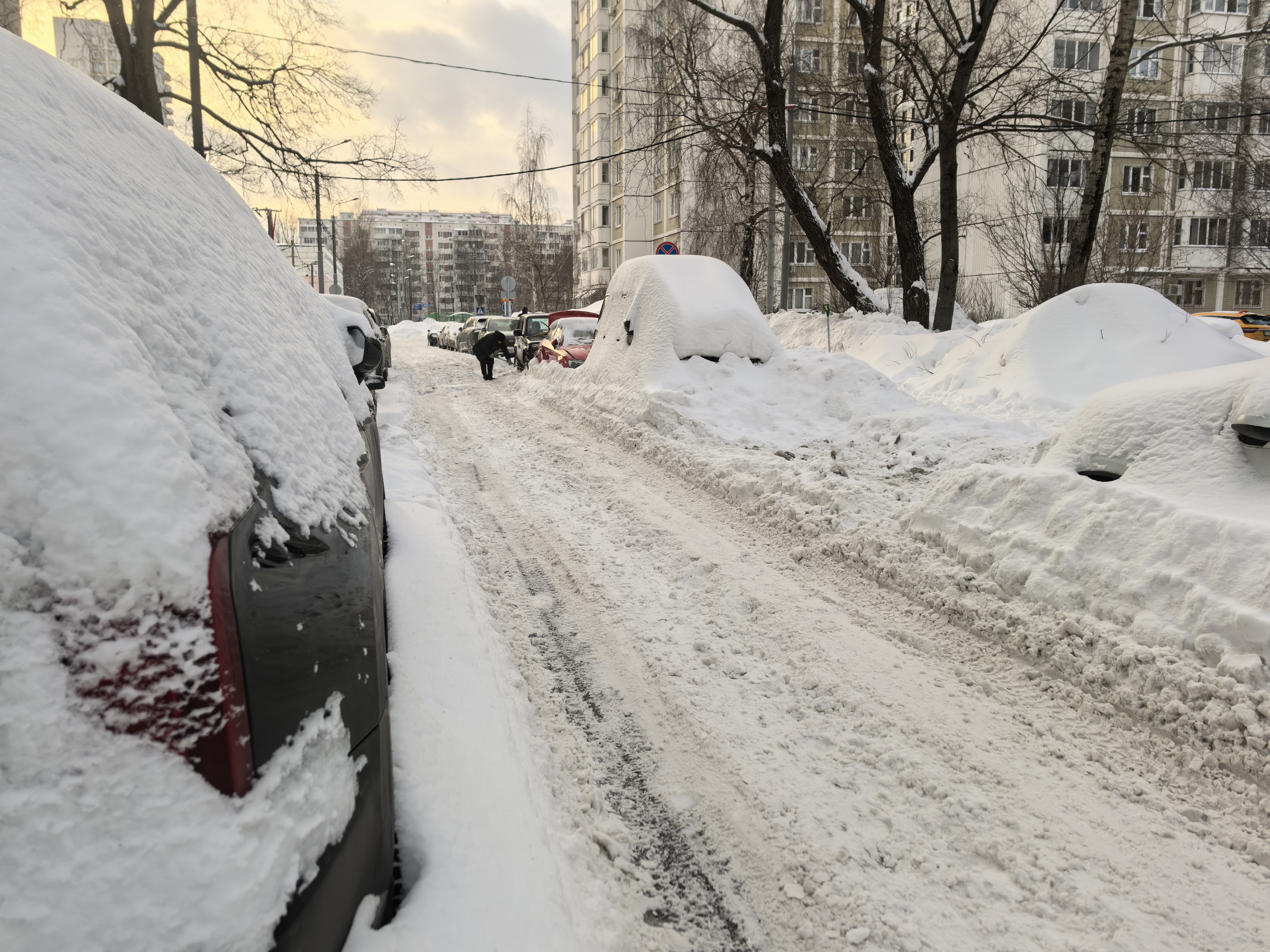 Дворовая территория с парковкой автомобилей, покрытых снегом. Проезжая часть узкая, заснеженная, без разметки. Видны жилые дома.