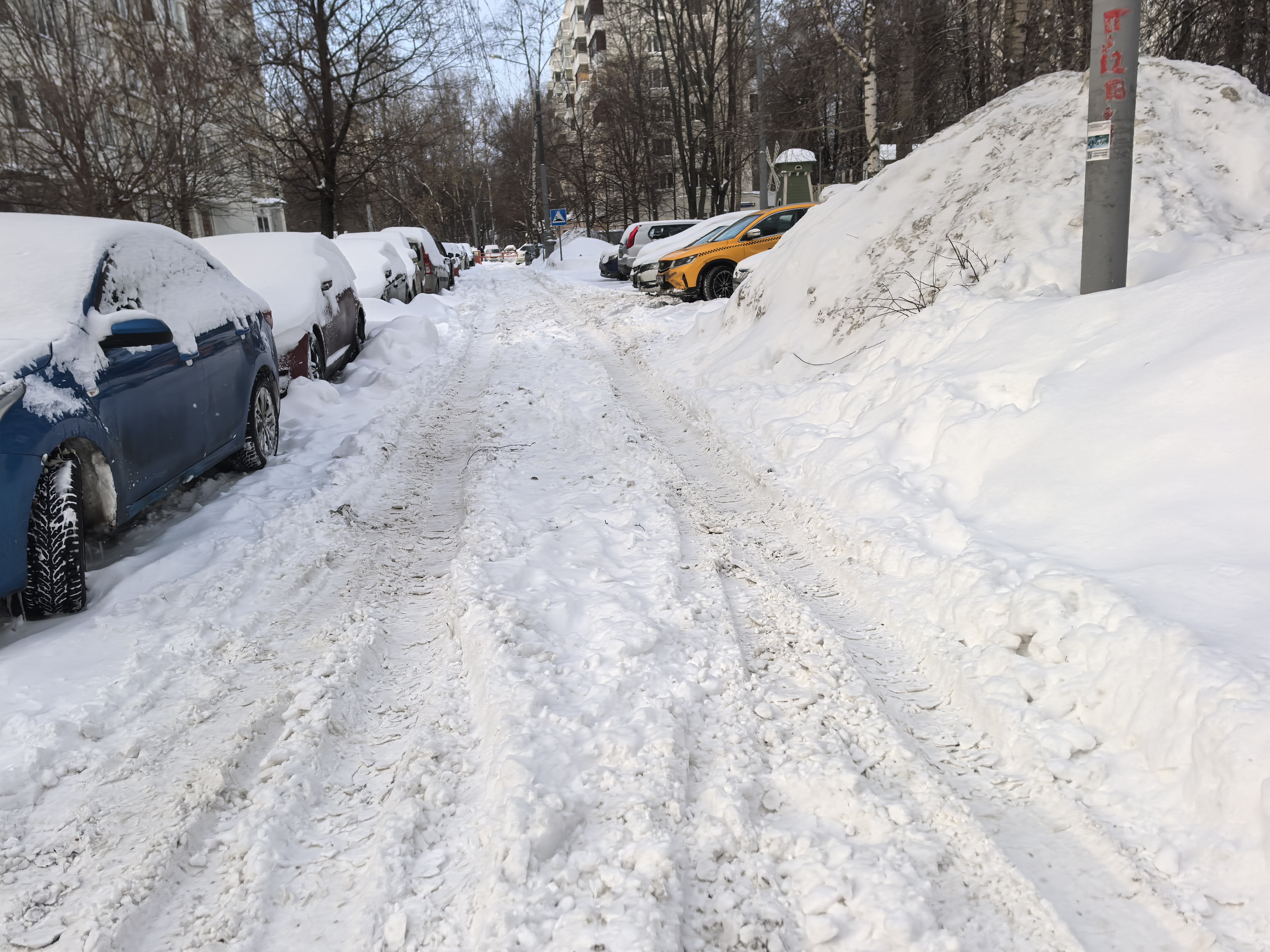 Участок городской дороги с пешеходным переходом, заснеженный, с припаркованными автомобилями по обеим сторонам.