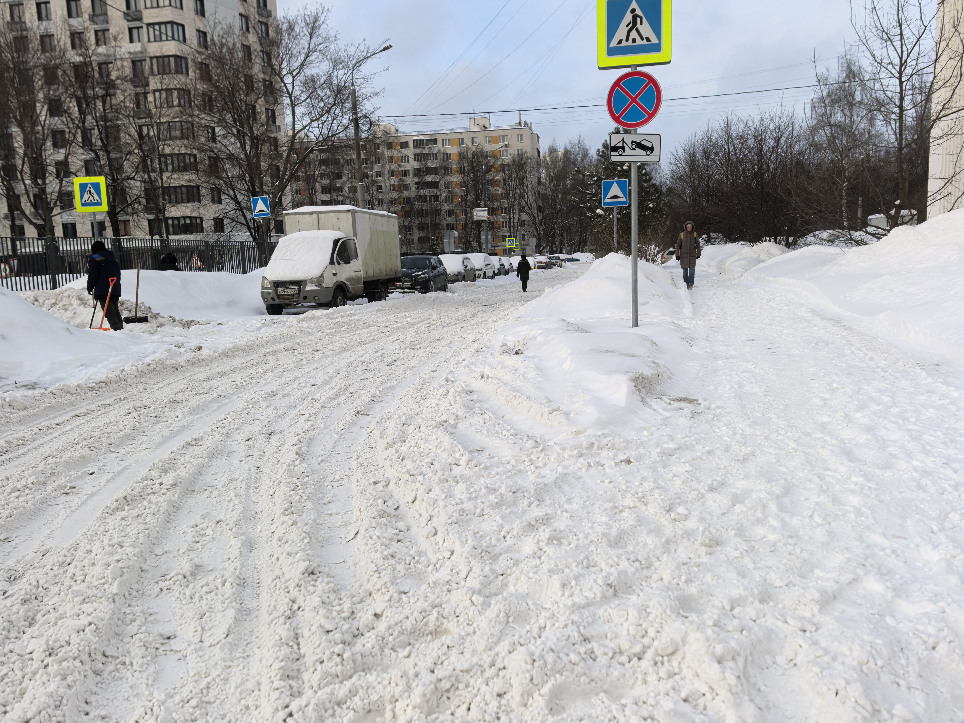Зимний городской участок дороги с пешеходным переходом, покрытым снегом. Видны знаки перехода, припаркованные автомобили и пешеходы.