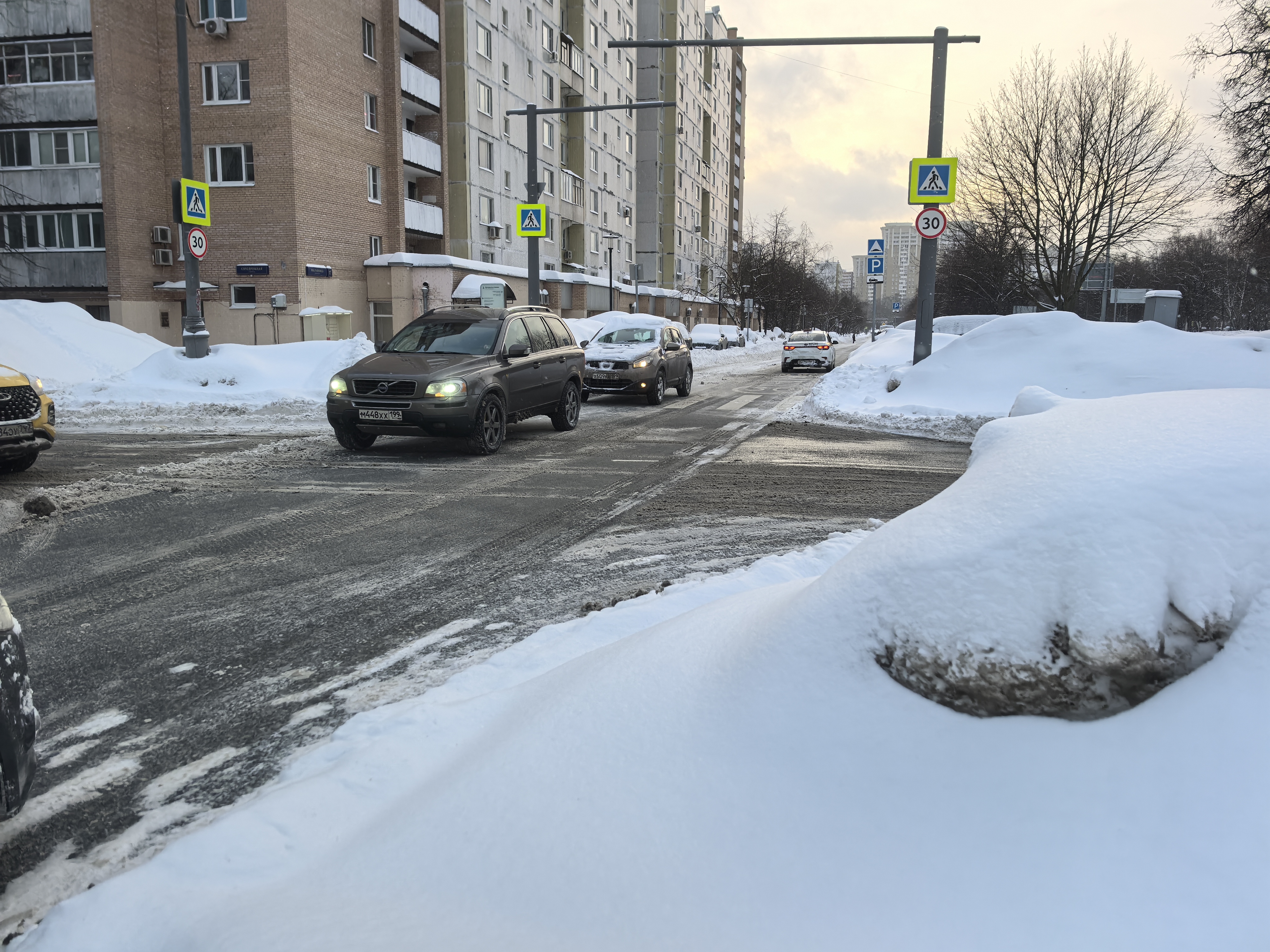 Участок городской дороги с пешеходным переходом, оформленным знаками и разметкой, в зимних условиях. На проезжей части видны движущиеся автомобили.