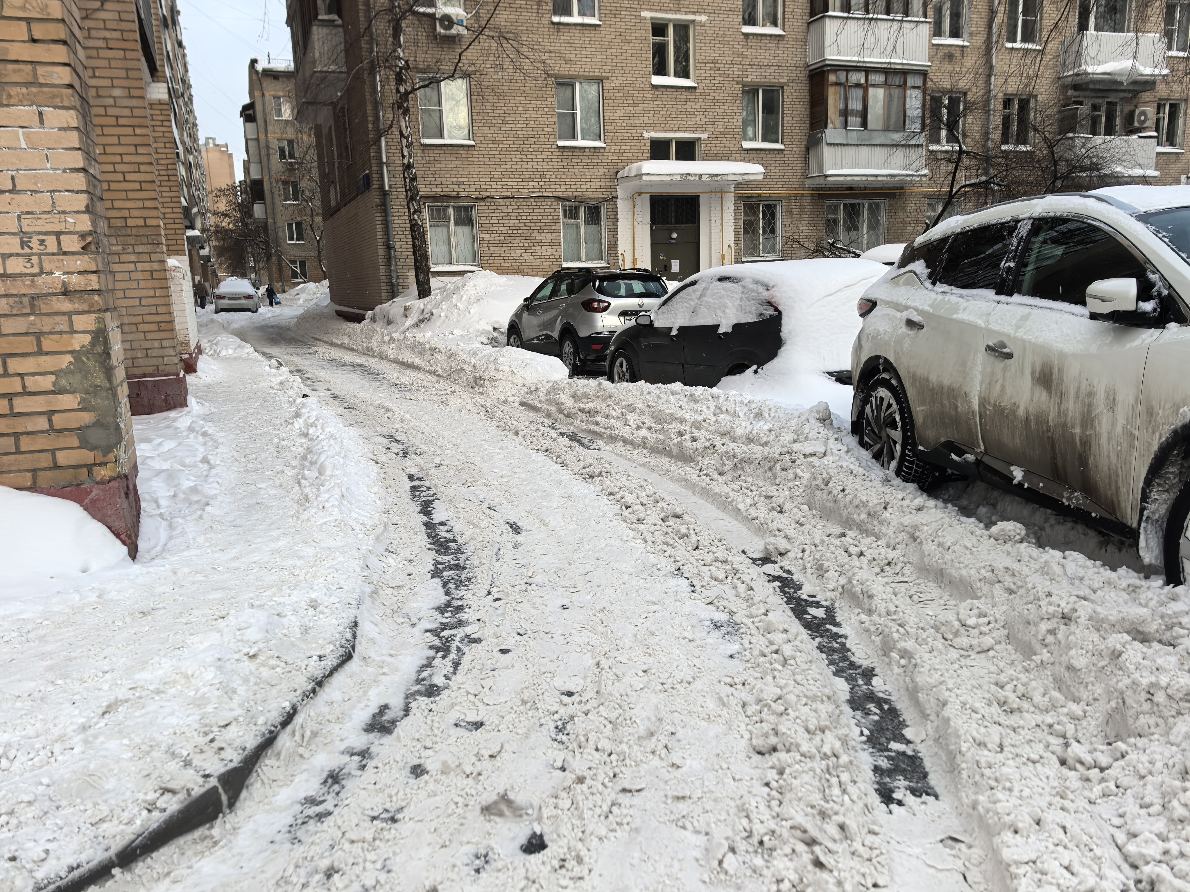 Дворовая территория с парковкой автомобилей вдоль домов, покрытая снегом и следами от шин.
