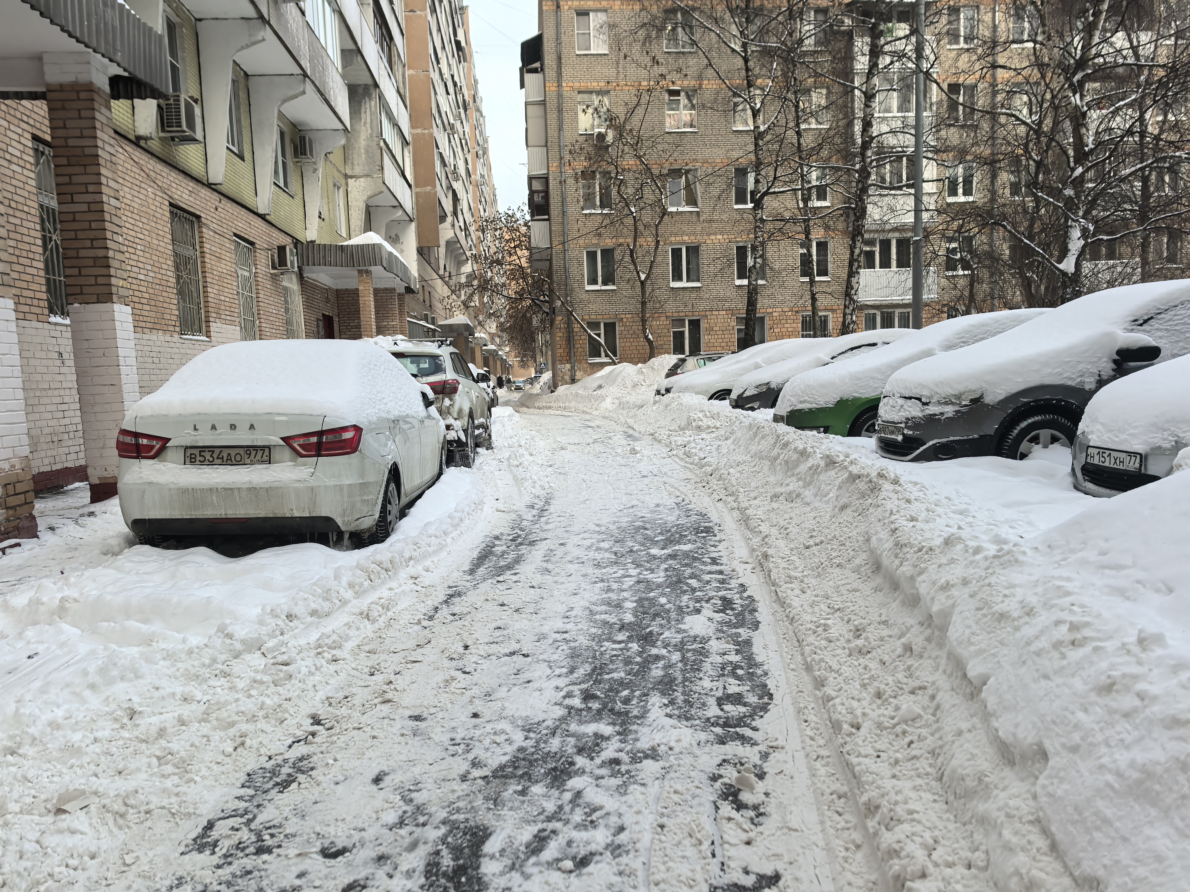 Дворовая территория между жилыми домами, покрытая снегом. Проезжая часть узкая, с припаркованными автомобилями по обеим сторонам.