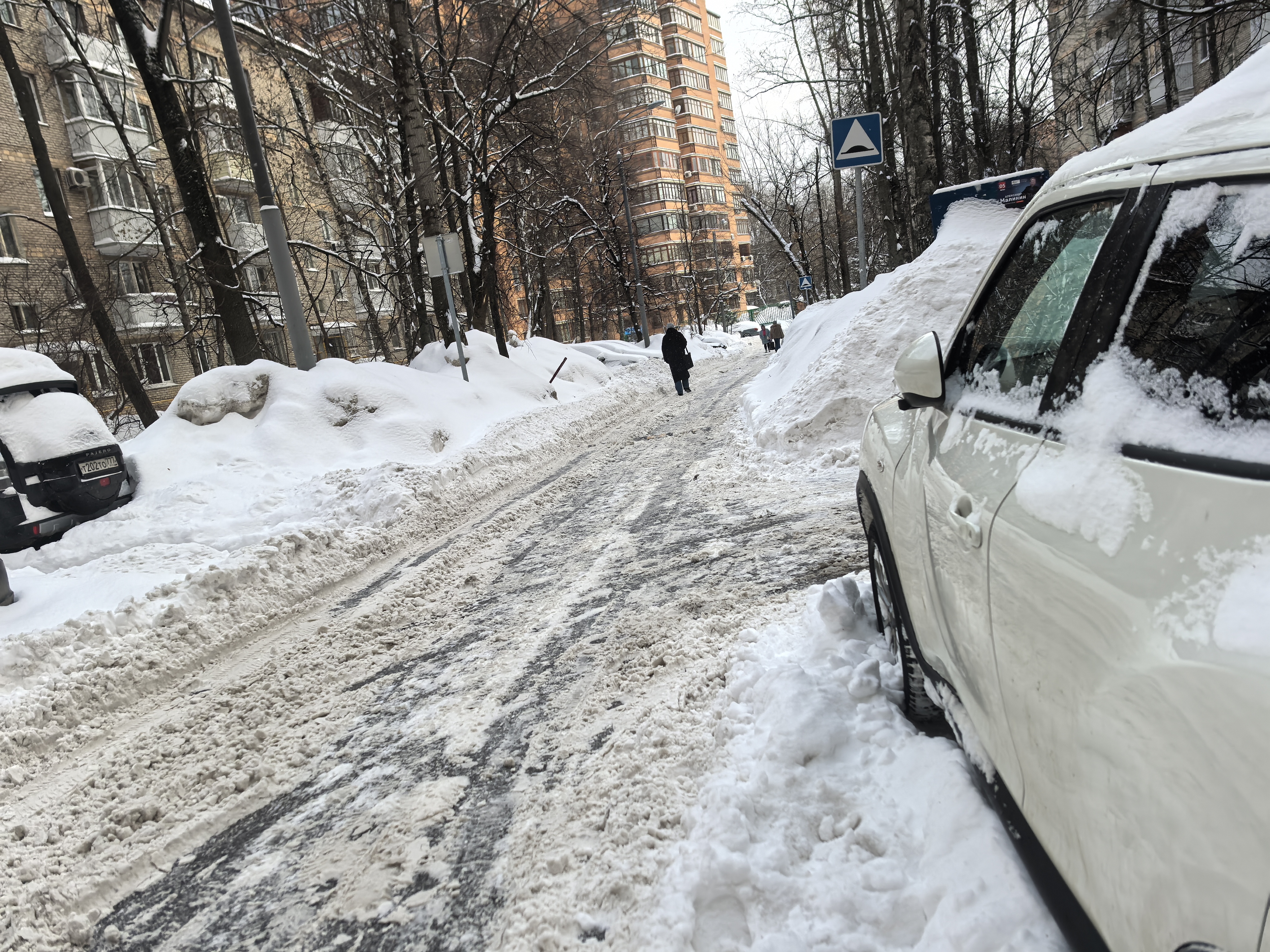 Дворовая территория, заснеженная дорога между жилыми домами, припаркованы автомобили, виден пешеход в центре проезжей части.