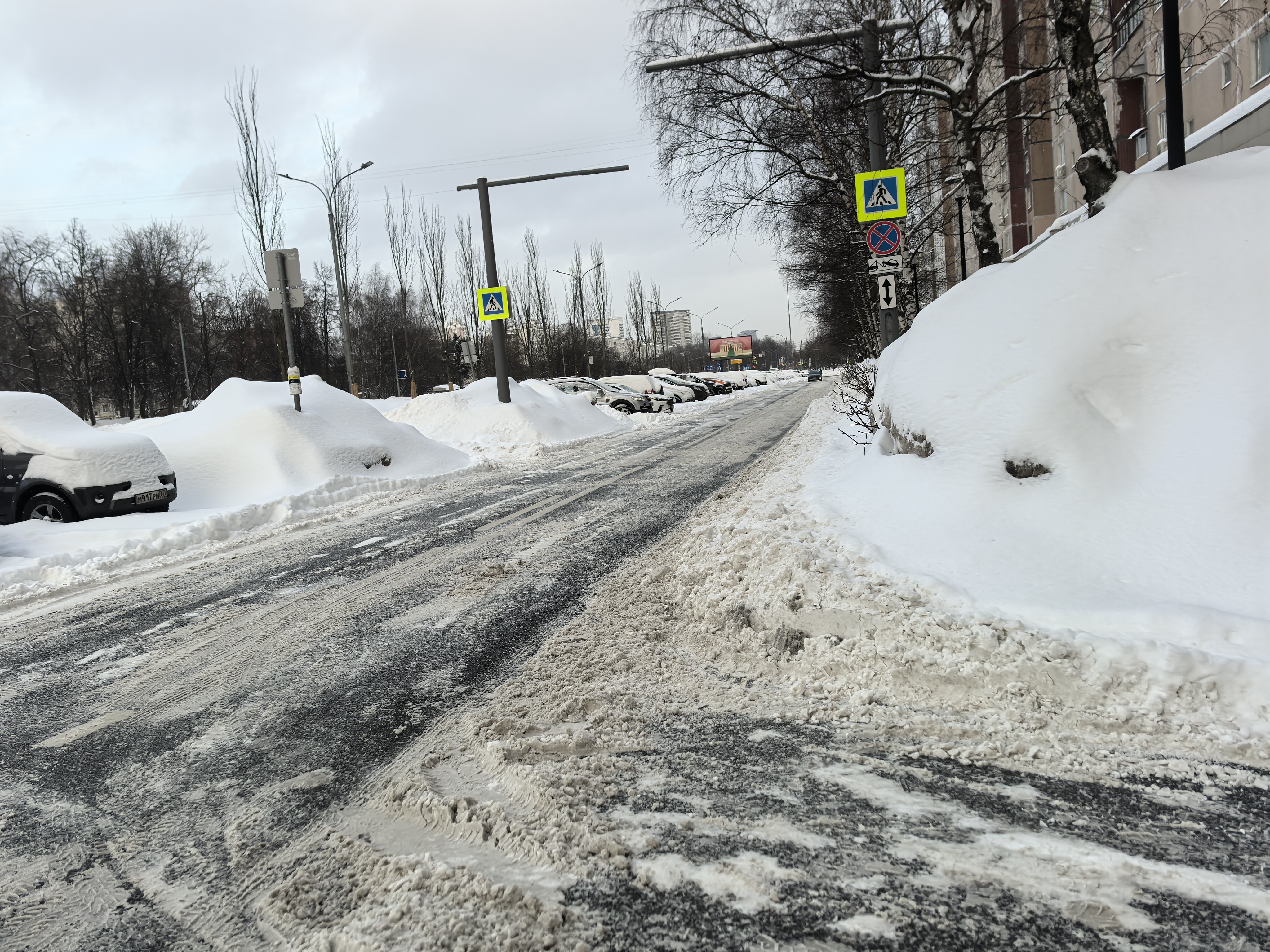 Участок дороги с пешеходным переходом, обозначенным знаками, в зимних условиях. Дорога покрыта снегом и наледью, видны припаркованные автомобили по обеим сторонам.