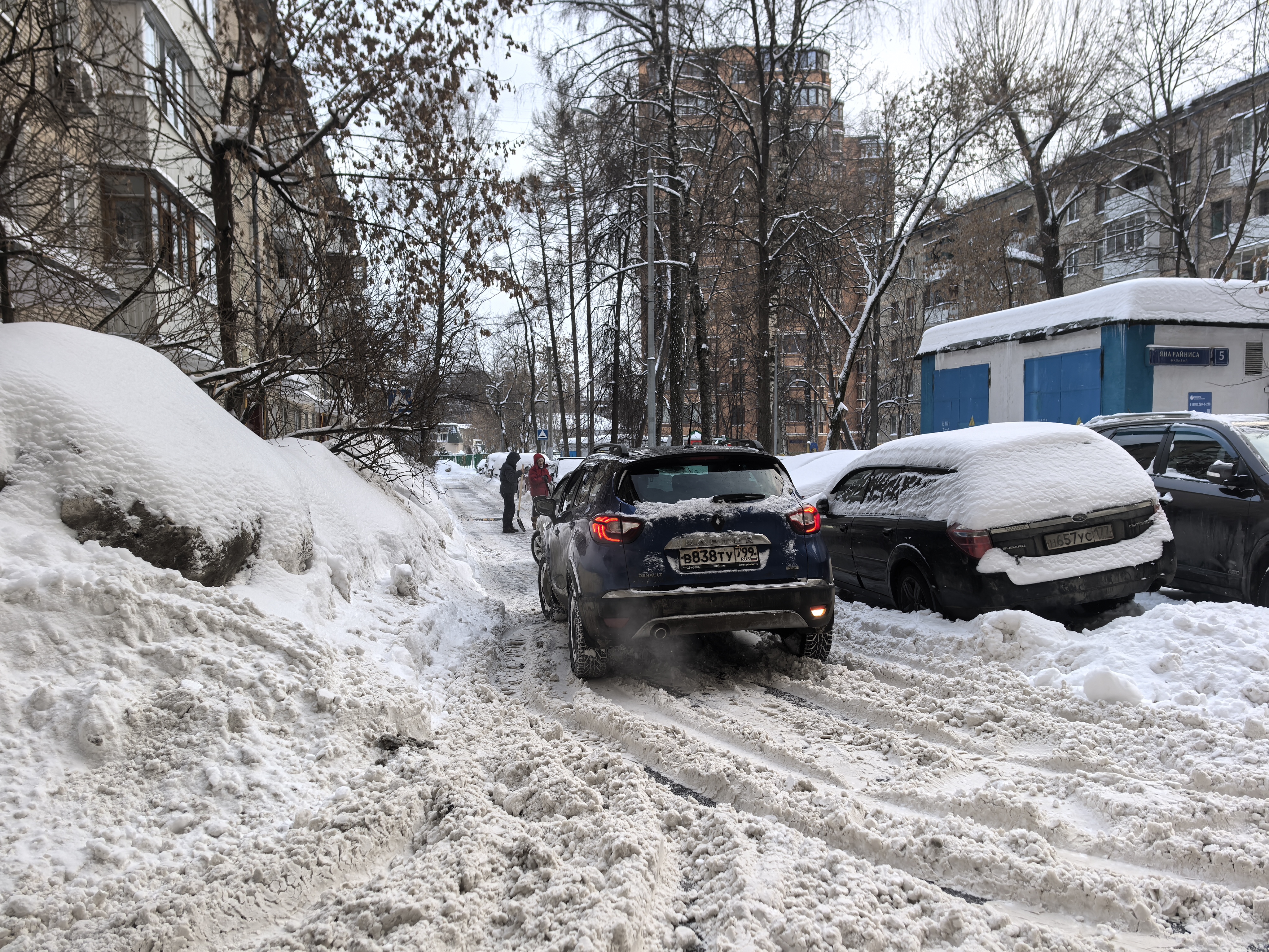 Дворовая территория с припаркованными автомобилями, покрытая снегом. Видны жилые дома и гаражи.