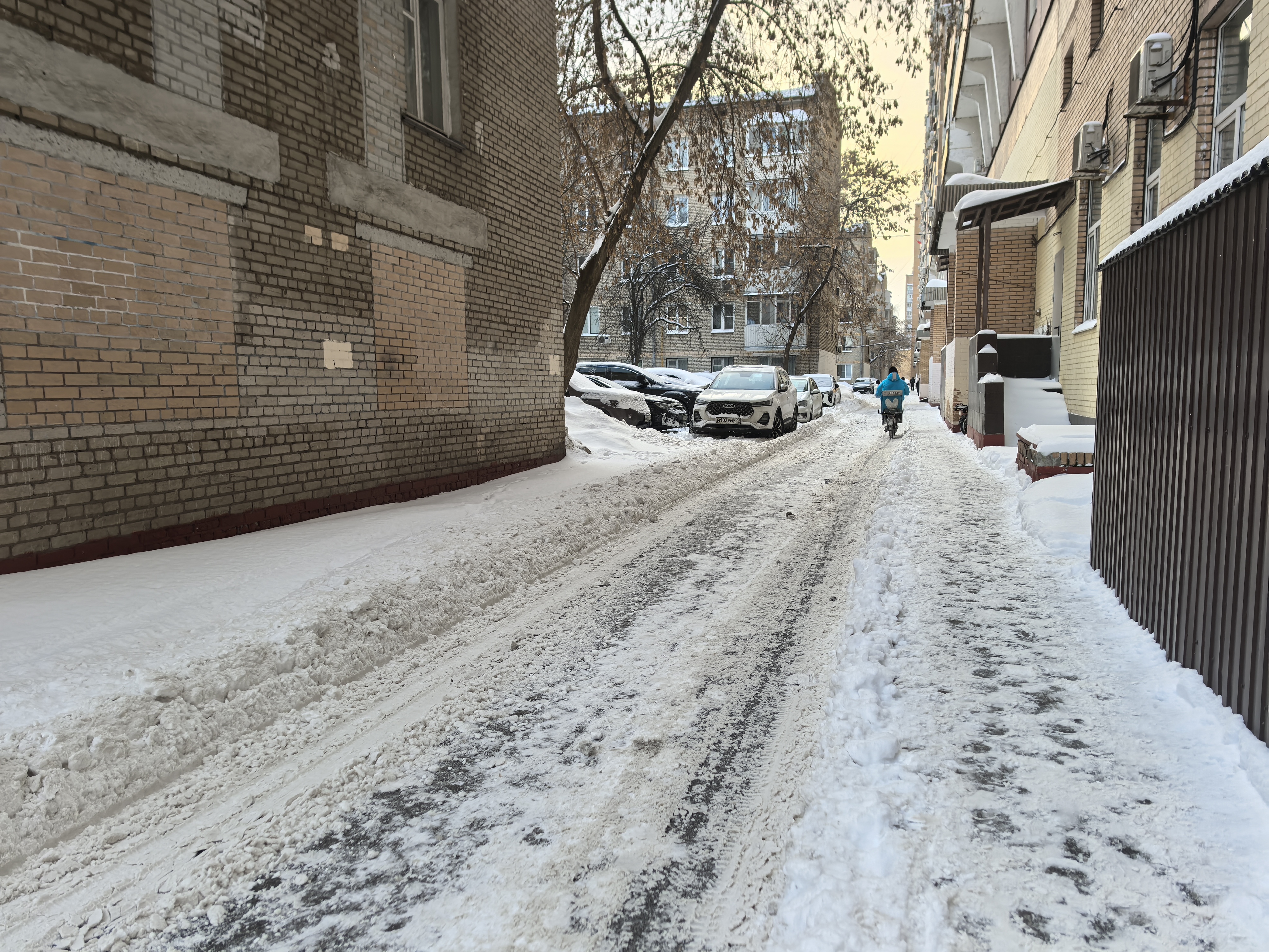 Дворовая территория между жилыми домами, покрытая снегом. Проезжая часть узкая, припаркованные автомобили занимают значительную часть пространства, сужая проезд.