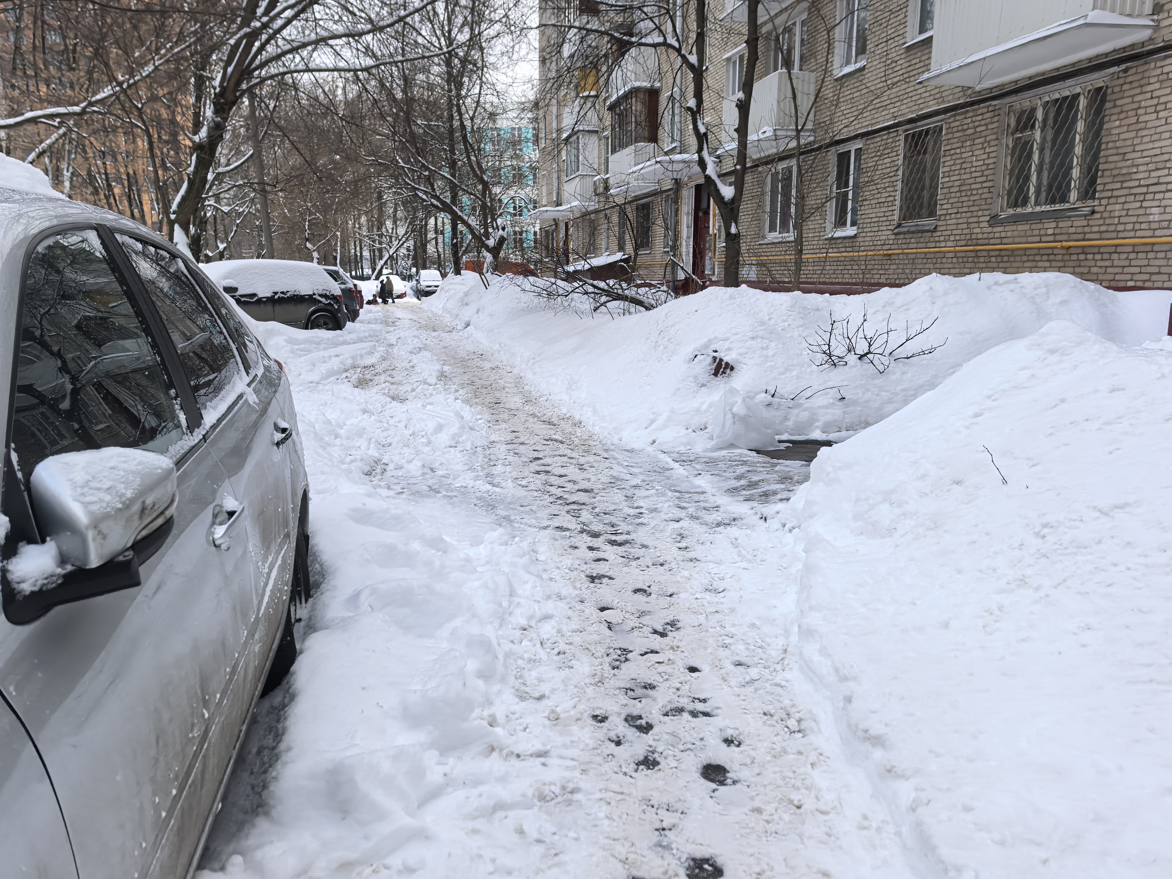 Дворовая территория с припаркованными автомобилями, покрытая снегом и наледью. Проход для пешеходов затруднен из-за высоких сугробов и узкой проезжей части.