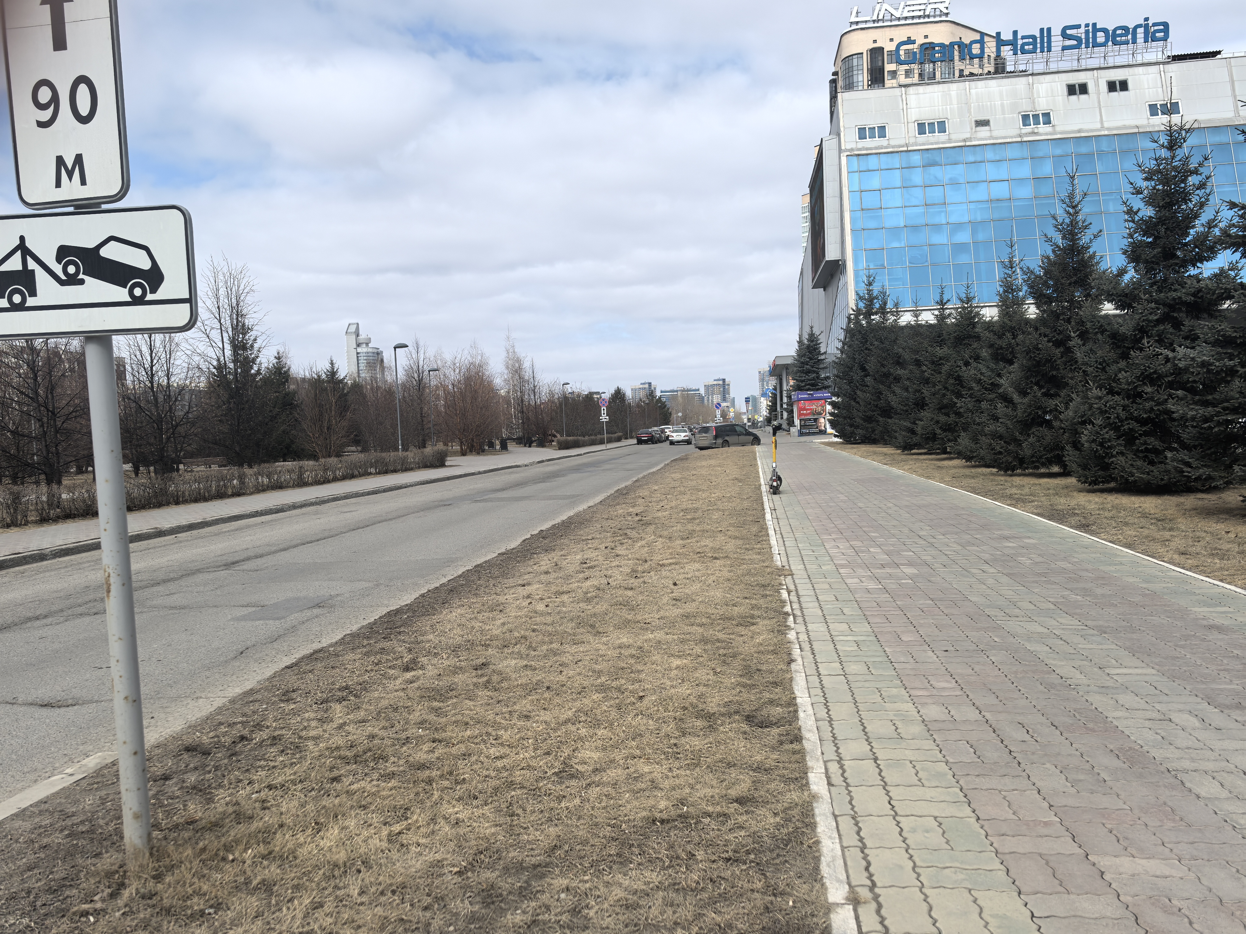 Проезжая часть без пешеходного перехода. Видна широкая проезжая часть, разделенная с тротуаром полосой сухой травы. На тротуаре уложена плитка, он ровный. Вдали видны автомобили, движущиеся по дороге. Знак эвакуации и табличка '90 м' указывают на приближе