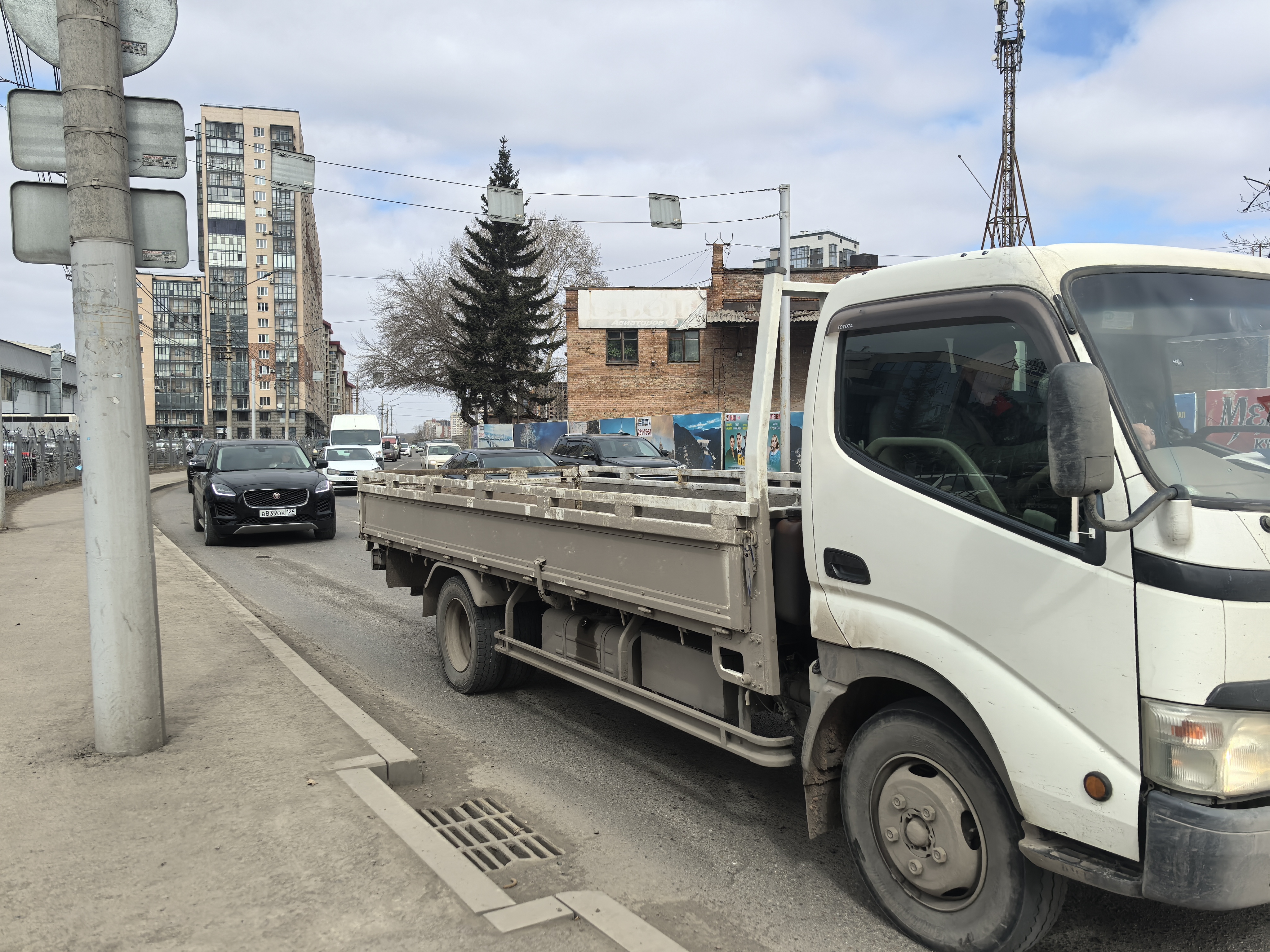 Участок городской дороги без пешеходного перехода. Видна проезжая часть с движущимся транспортом, тротуар слева отделен бордюром. На заднем плане видны жилые дома и коммерческие здания.