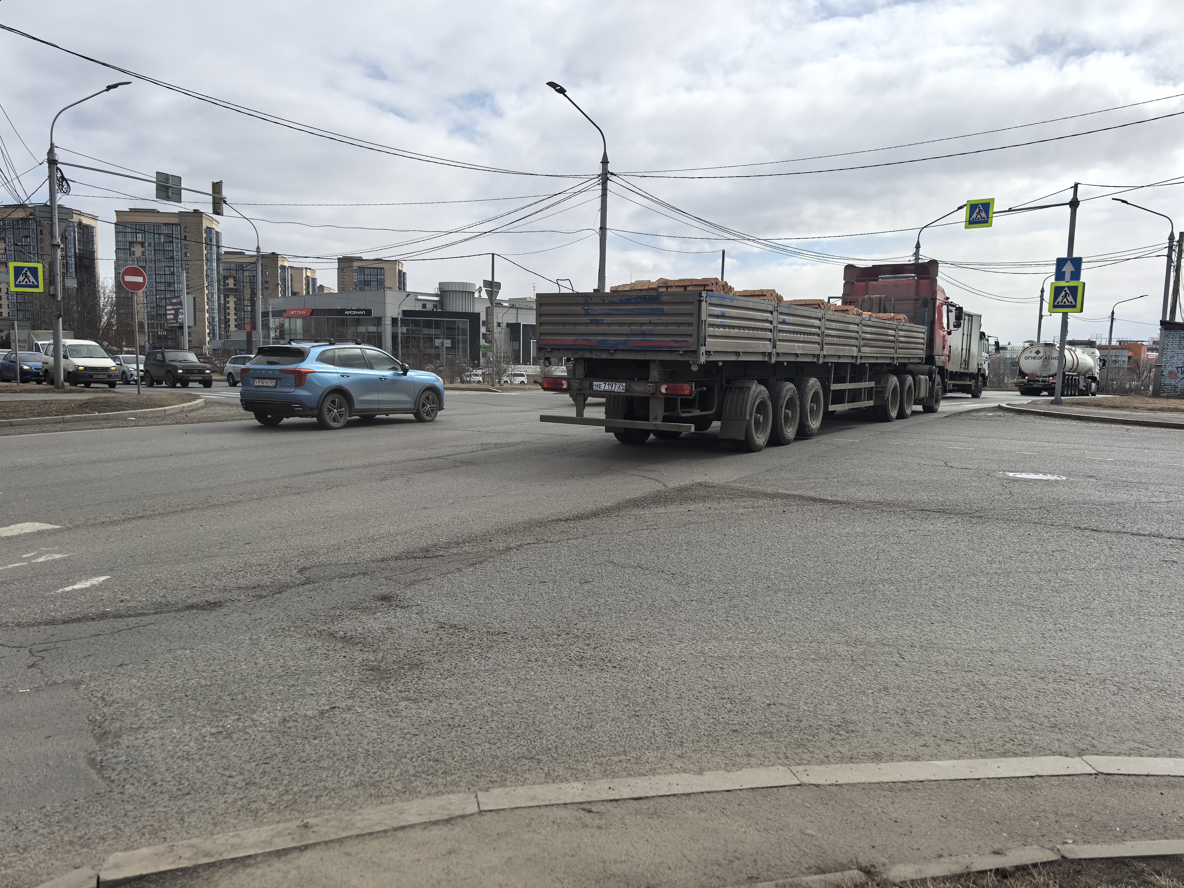 Широкая городская дорога с организованным пешеходным переходом, обозначенным знаками и разметкой. Видны движущиеся транспортные средства и припаркованные авто на обочине.