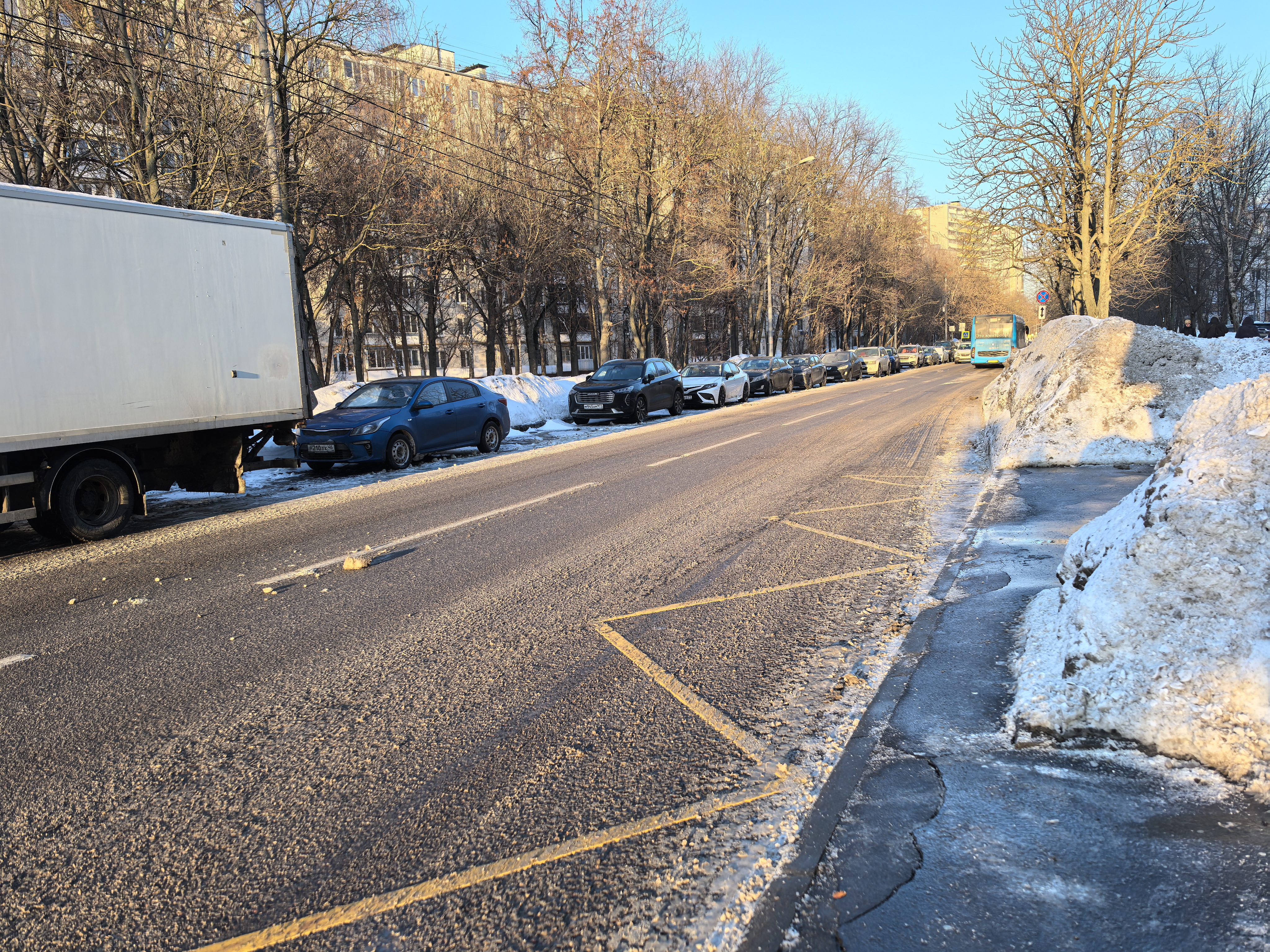 Проезжая часть без пешеходного перехода. Видна припаркованная техника и ряд автомобилей, тротуар частично засыпан снегом.