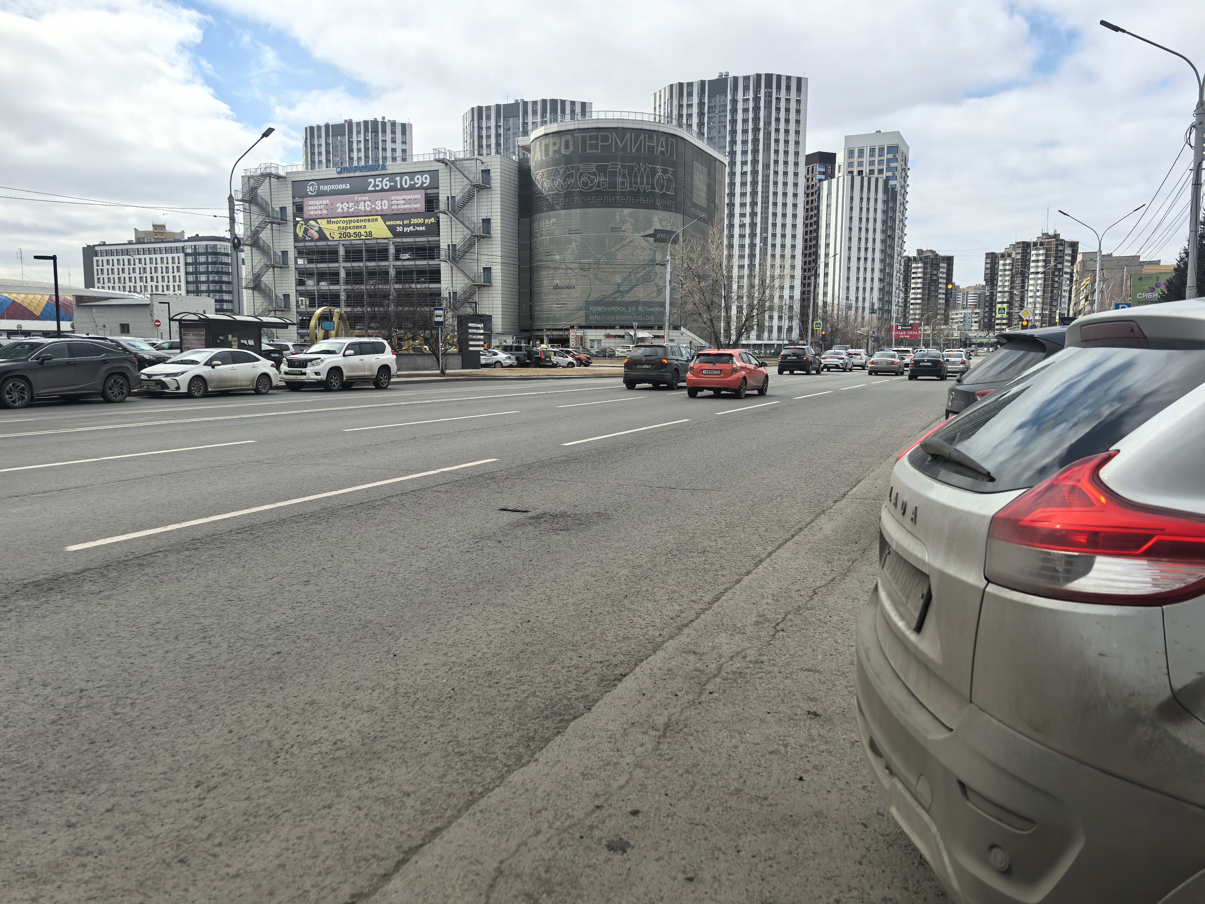Широкая городская проезжая часть с интенсивным движением, без пешеходного перехода в зоне видимости. Справа видна припаркованная машина, слева — припаркованные авто и остановка. Тротуары есть, но переход отсутствует.