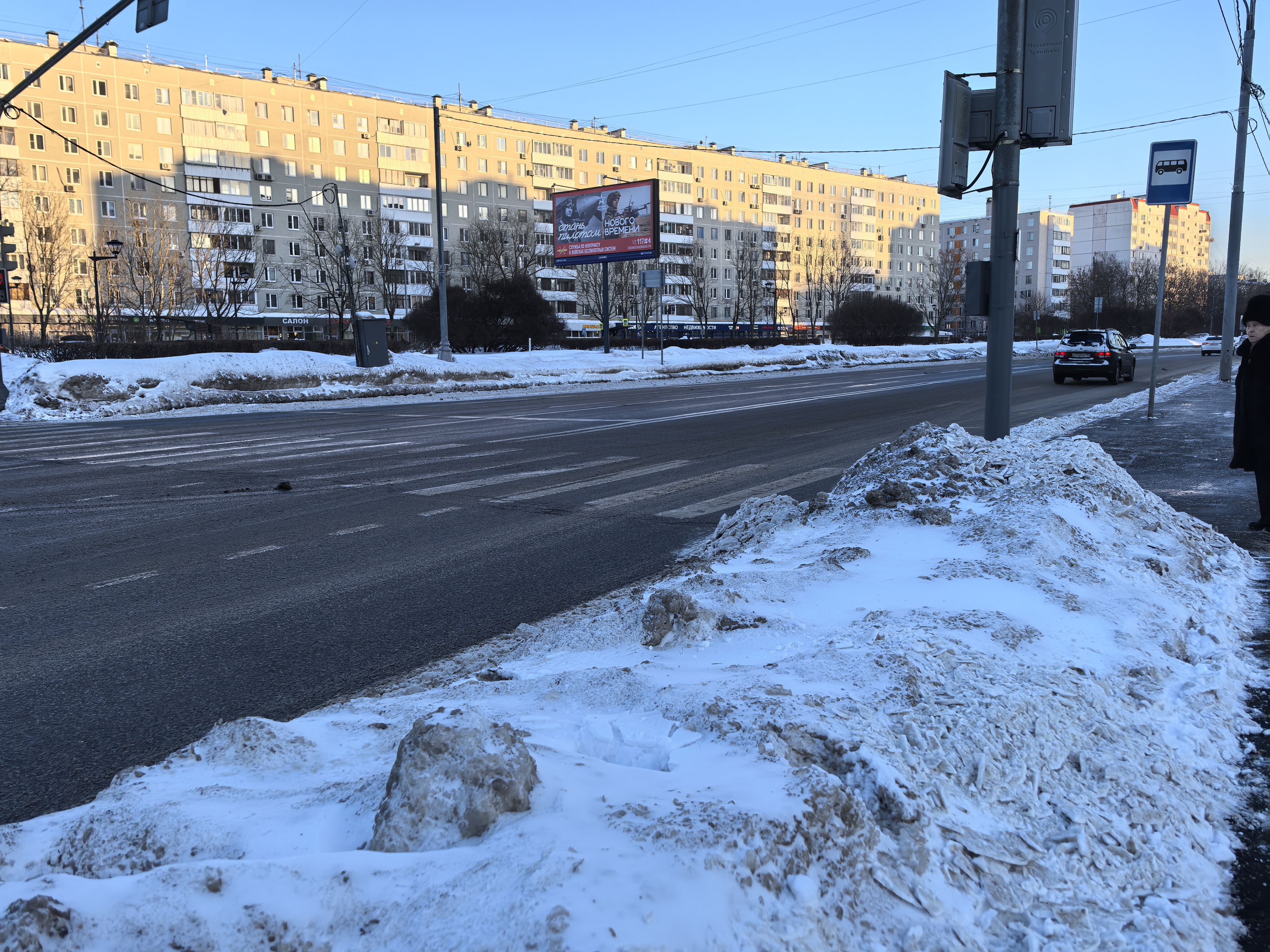 Широкая городская дорога с пешеходным переходом (зебра), зимнее время, на обочине большие сугробы, видны многоэтажные жилые дома.